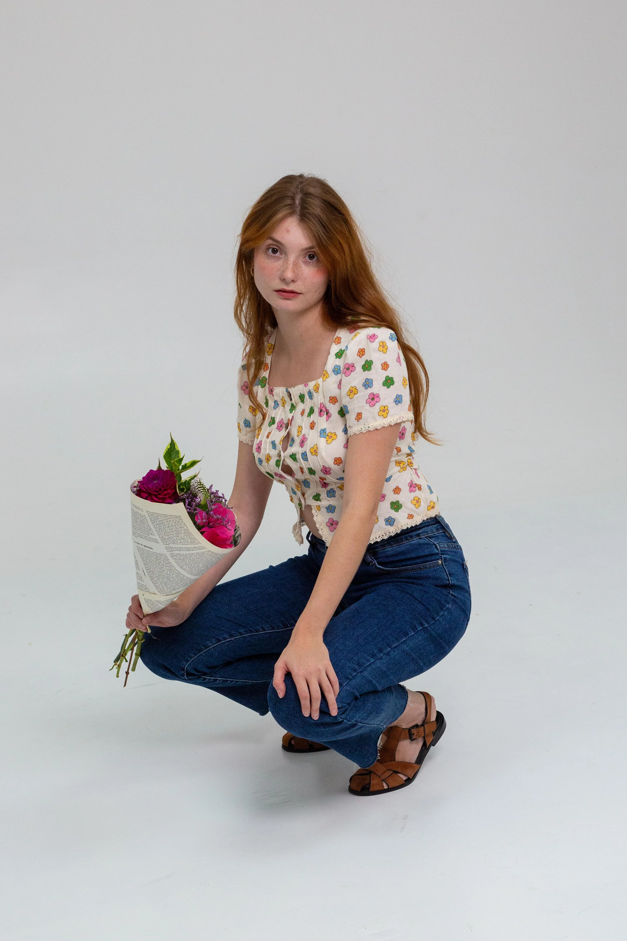 Jeune femme aux cheveux roux, portant un chemisier à fleurs, tenant un bouquet de fleurs dans un magazine, en position accroupie sur un fond blanc.
