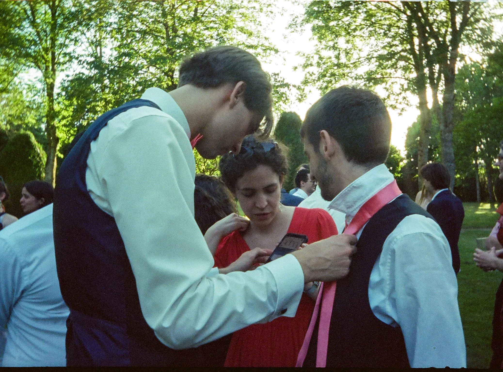 Un groupe de jeunes adultes en costume et habillés élégamment lors d'un mariage en extérieur,  vérifient comment mettre une cravate sur un téléphone portable, en arrière-plan la lumière passe à travers les arbres.