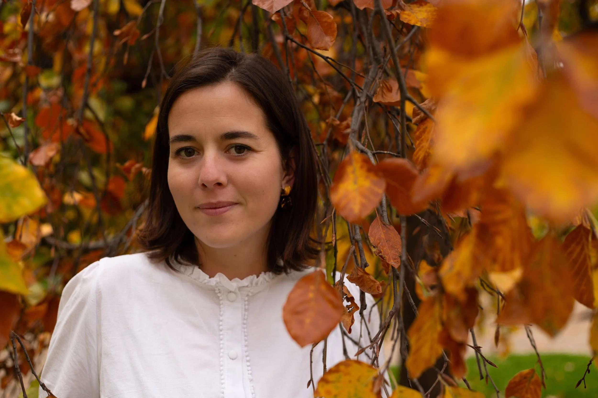 Une femme avec les cheveux bruns courts portant une chemise blanche se tient près d'une haie d'automne avec des feuilles orange et marron.