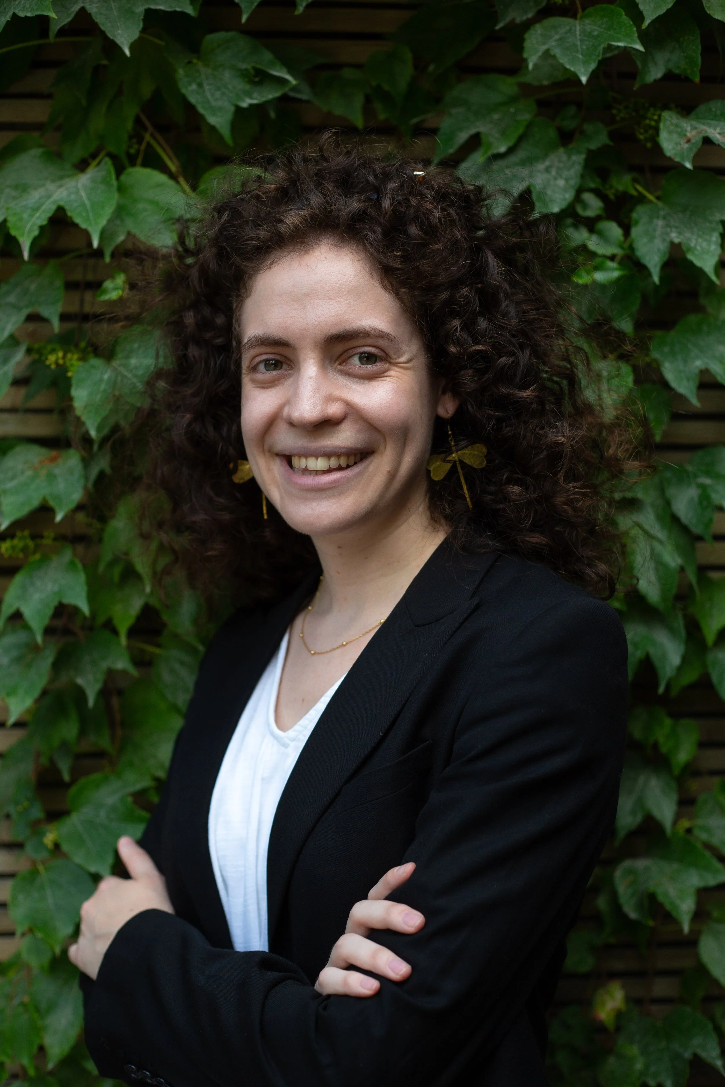 Une femme souriante en tenue professionnelle, portant des boucles d'oreilles en forme de papillon et une chaîne délicate, se tient devant un mur recouvert de feuilles de lierre vert.