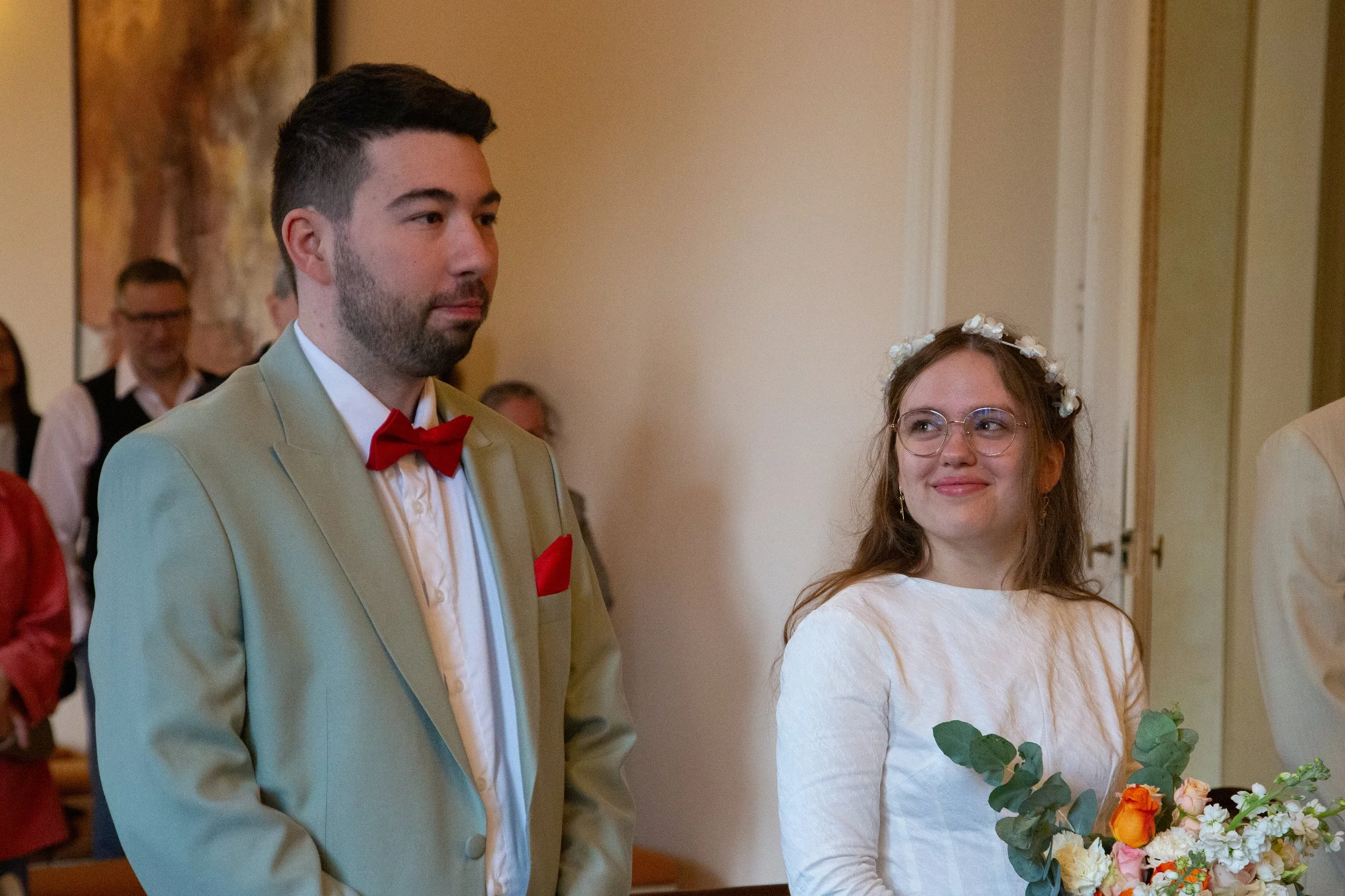 Un homme et une femme lors de leur mariage, l'homme en costume verte avec un nœud papillon corail et la femme en robe blanche avec une couronne de fleurs, tenant un bouquet de fleurs.