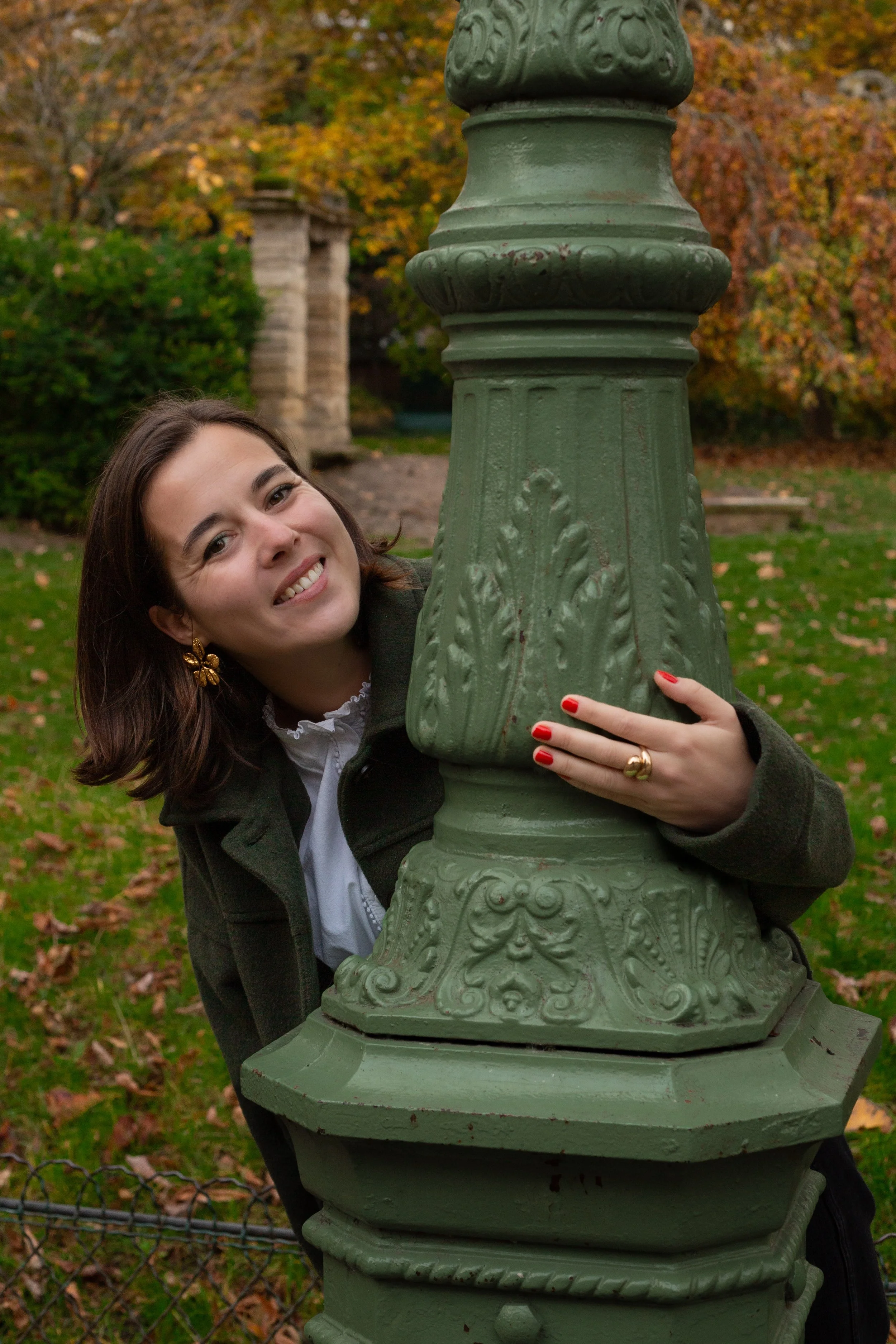Une femme souriante en automne, à côté d'une colonne décorative verte dans un parc avec des arbres aux feuilles orange, jaune et marron.