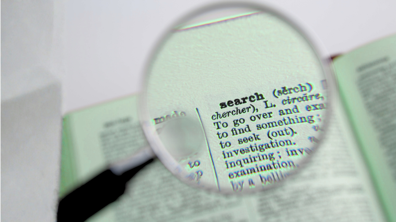 A dictionary opened to the word 'search' with a magnifying glass focusing on the definition, which describes searching as looking over and examining to find something.