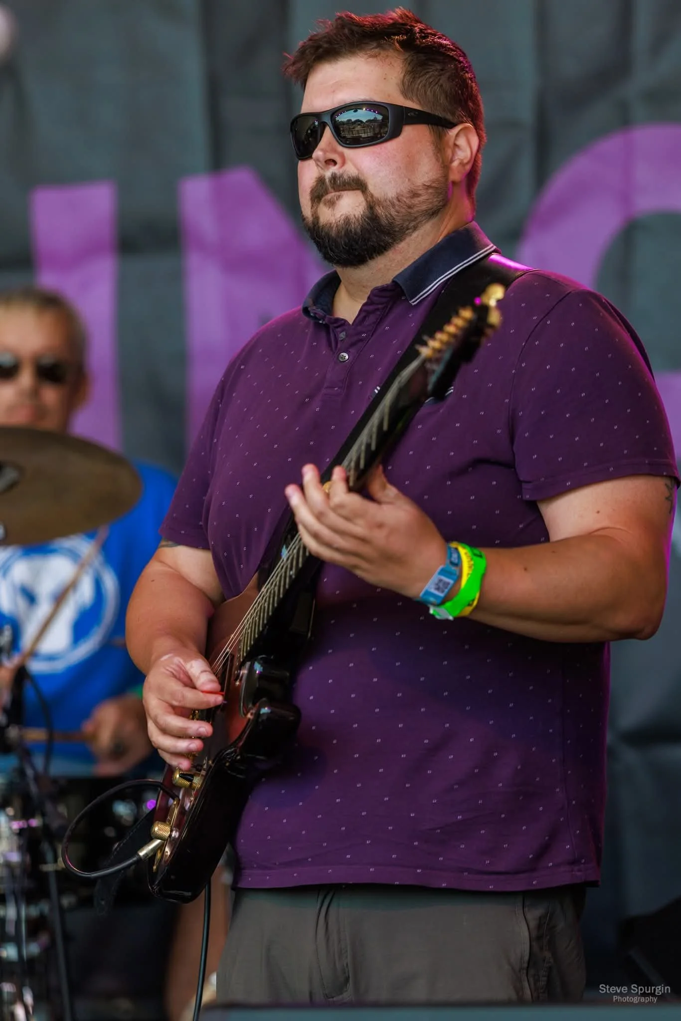 Jay in sunglasses playing an electric guitar during a performance at Victorious Festival 2025, wearing a purple shirt with small blue dots.