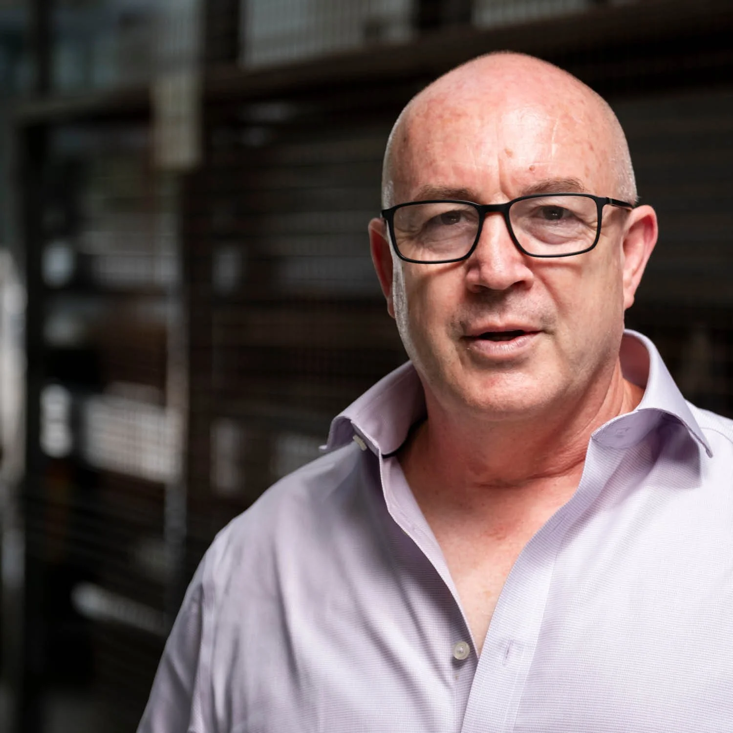 Close-up of a bald man with glasses wearing a light purple dress shirt, indoors with a blurred background of shelves.