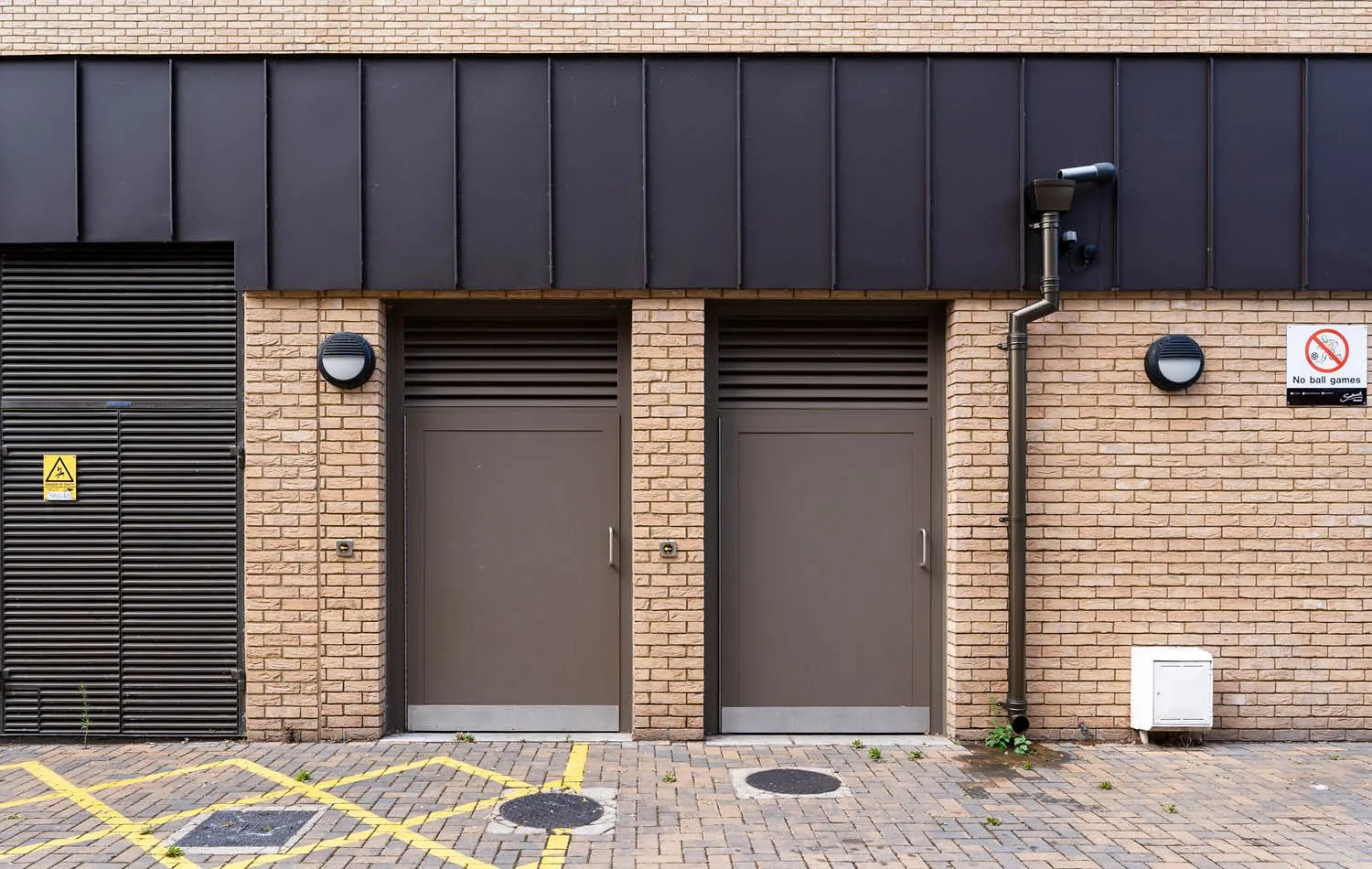 Brick building exterior with two grey doors, black vent covers, two wall-mounted lights, a drainage pipe, and signs indicating no ball games and caution for electrical equipment. The ground has yellow parking lines and small patches of weeds.