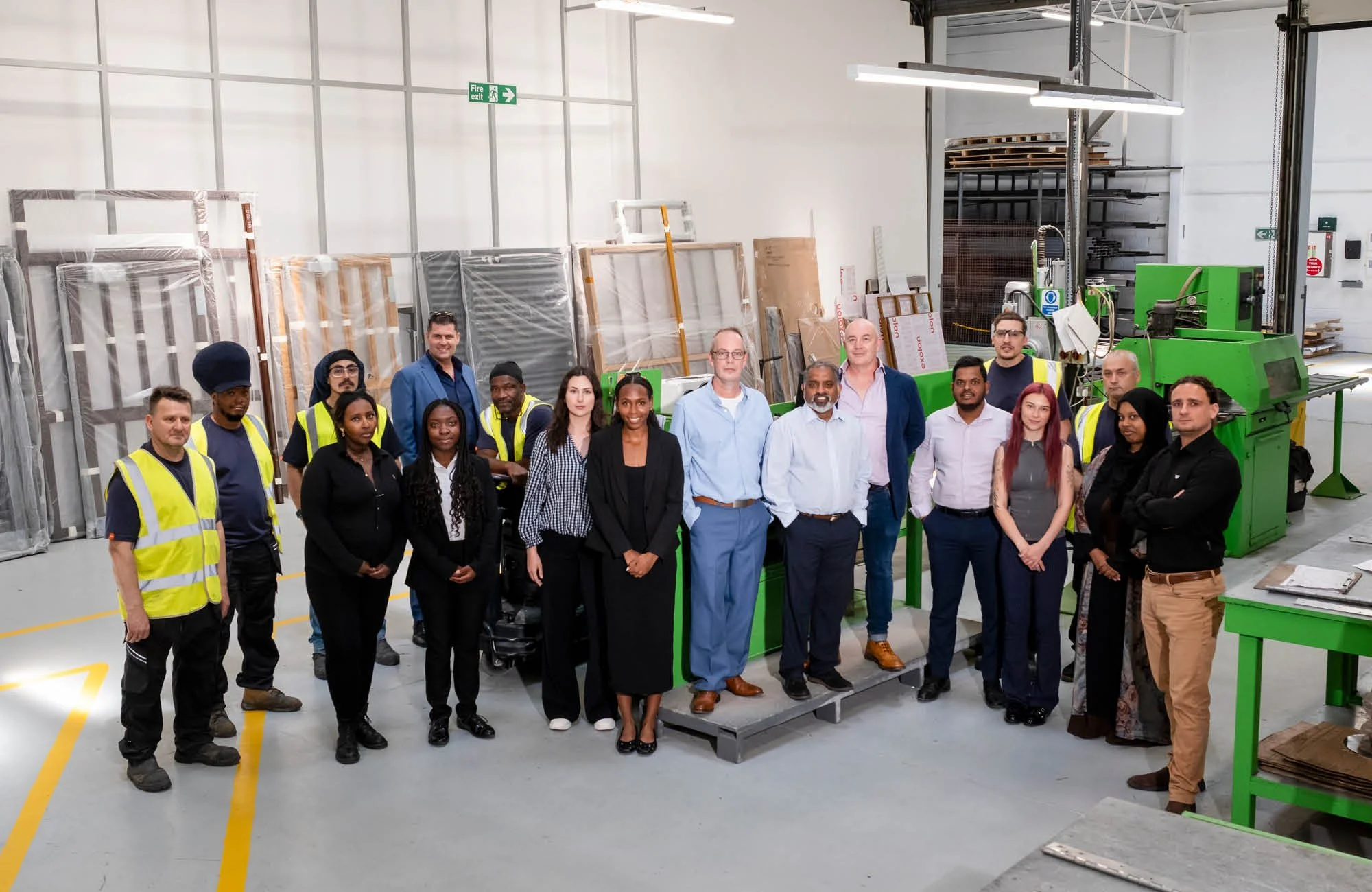 Group of diverse professionals standing together inside a manufacturing or industrial facility, surrounded by machinery, equipment, and storage racks.