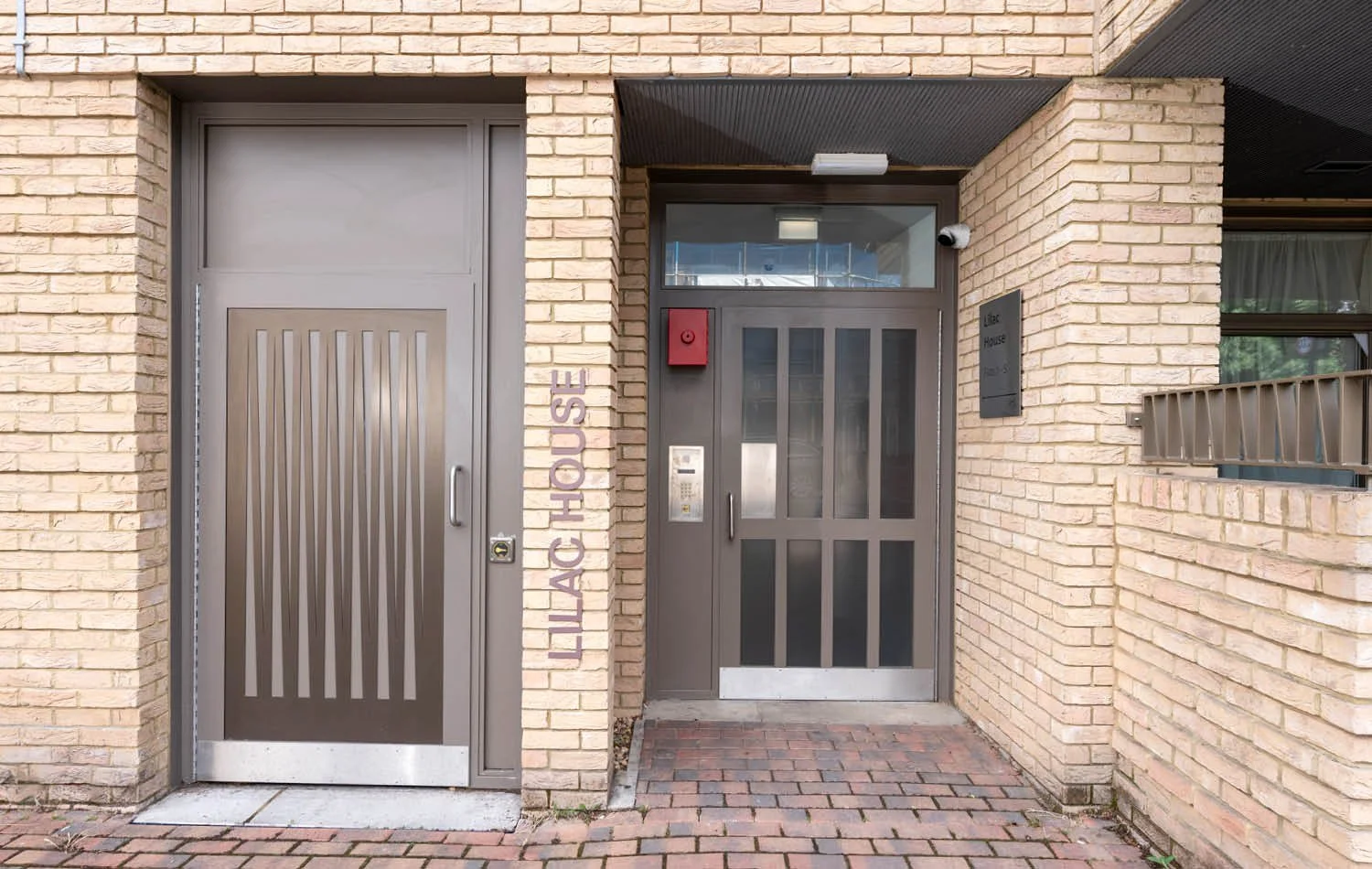 Brick building entrance with two gray doors, one labeled "LILAC HOUSE," with a red box, intercom, and security camera.