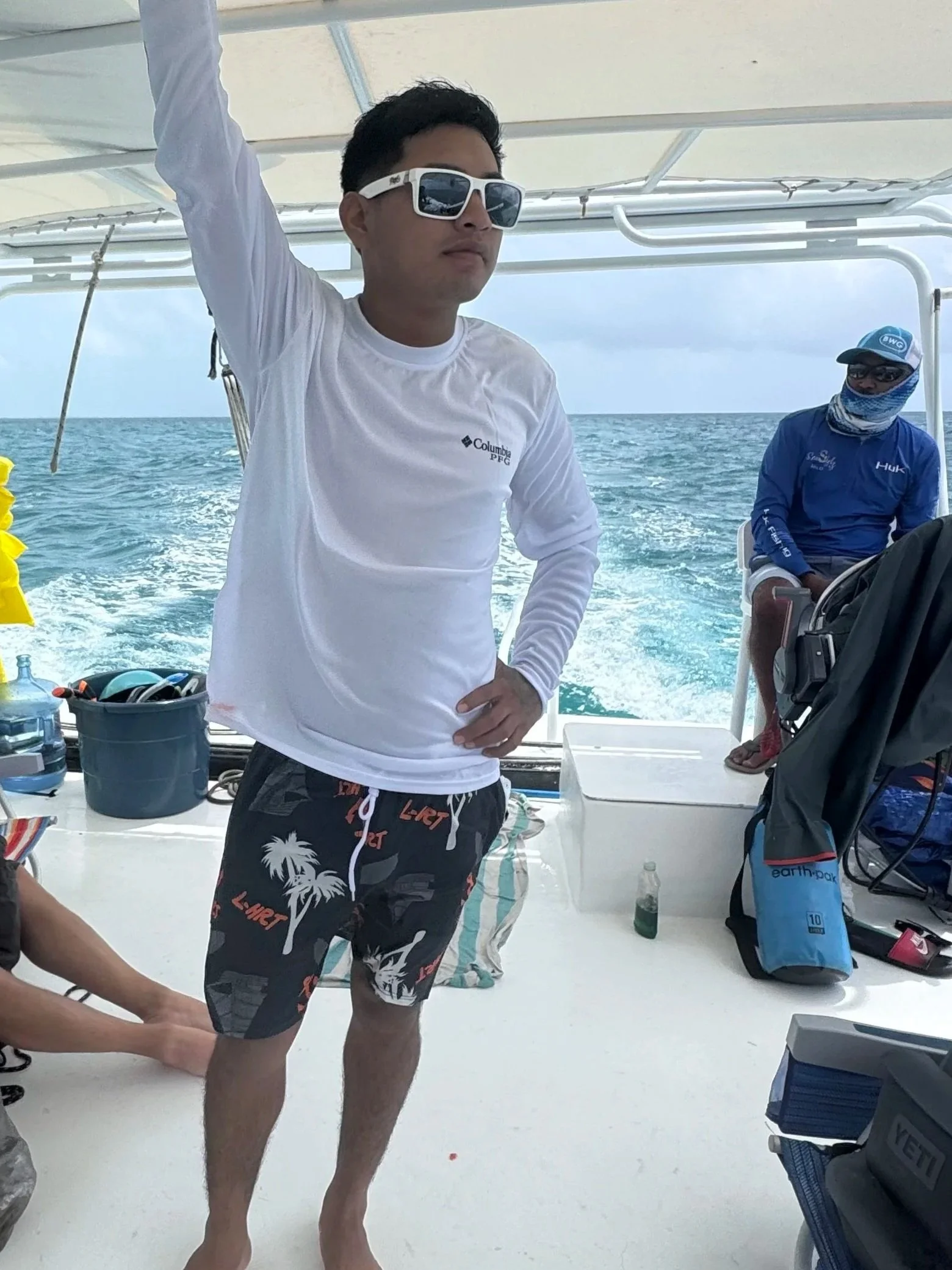 A young man standing on a boat, wearing a white long-sleeve shirt, tropical print shorts, and sunglasses, with the ocean in the background.