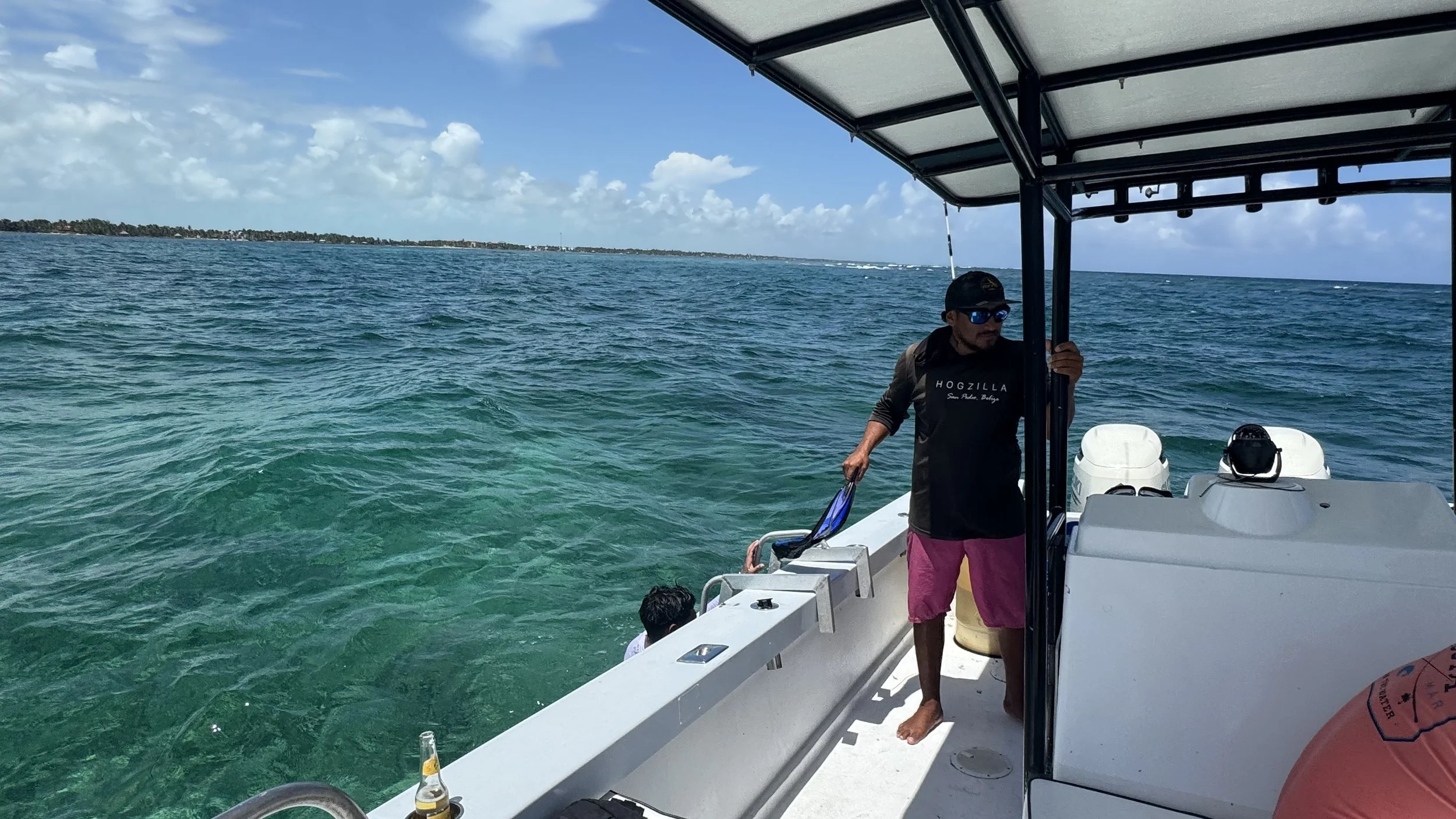 A man with sunglasses and a hat on a boat in the open ocean wearing pink shorts and a black shirt, with the shoreline visible in the distance under partly cloudy skies.