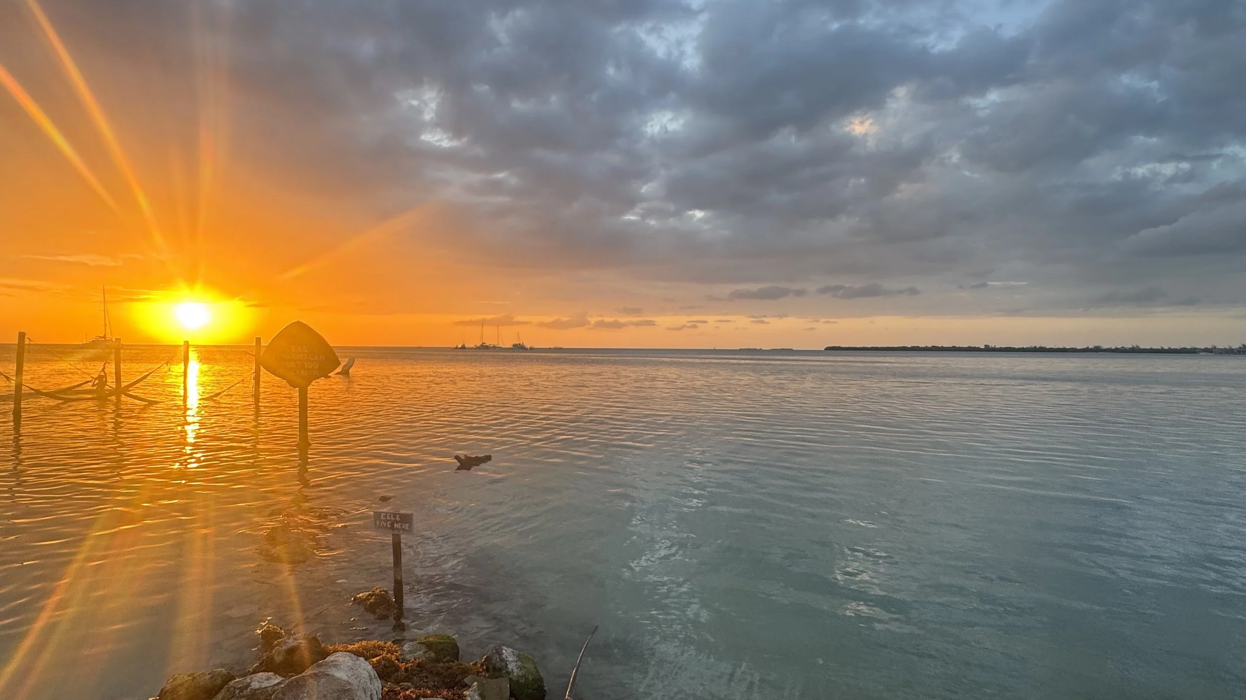 A sunset over a body of water with clouds in the sky. The sun is near the horizon, casting a warm orange glow. There are some boats and a sign in the water, with rocks along the shoreline.