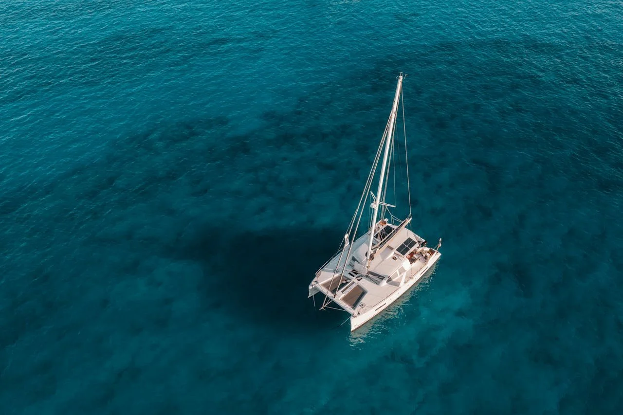 An aerial view of a white sailboat floating on clear blue ocean water.