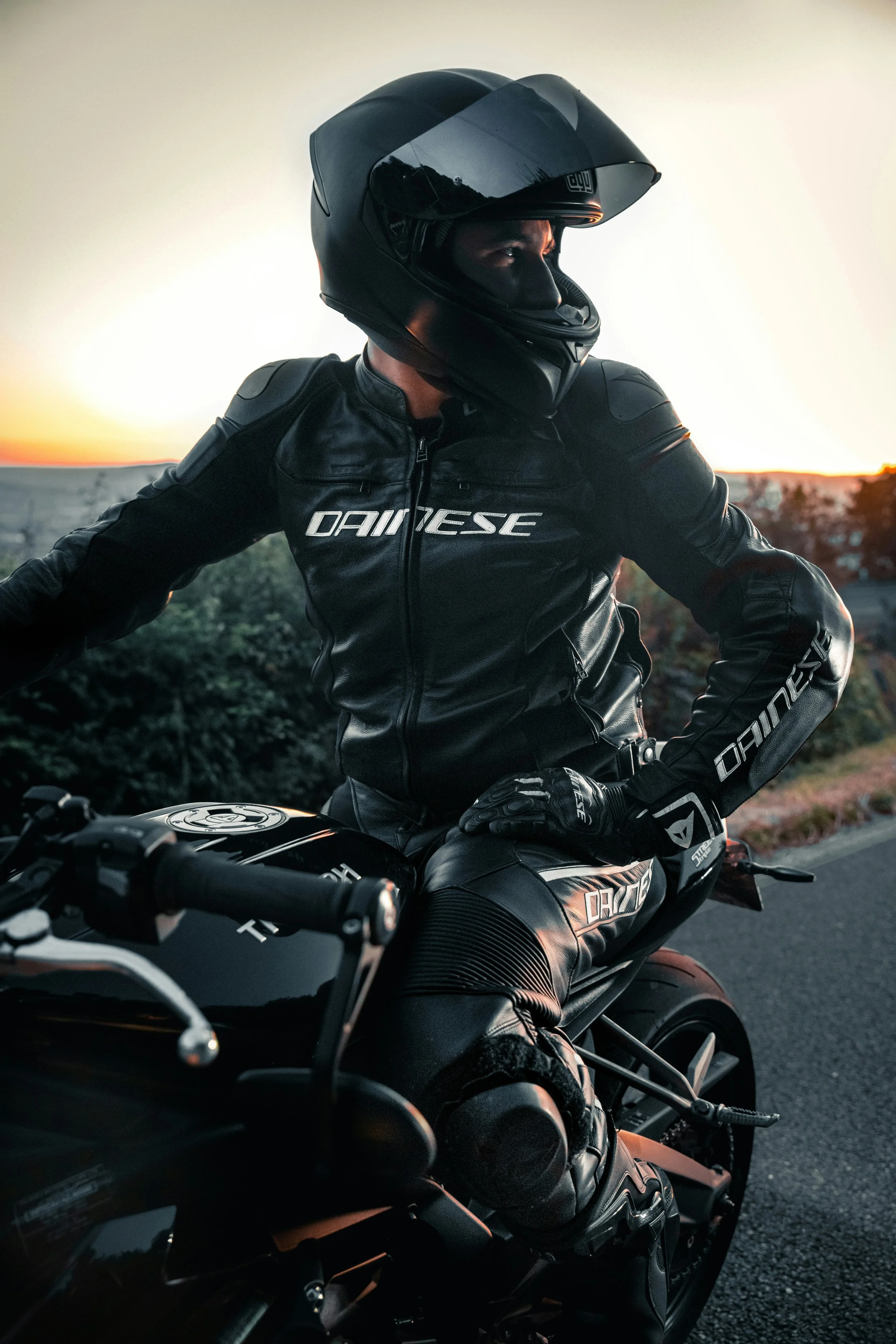 Pessoa vestida com roupa de proteção para moto, usando capacete, diante do pôr do sol, ao lado de uma motocicleta preta.