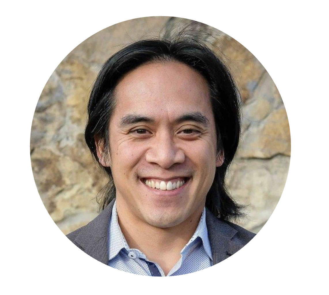 A smiling man with dark hair, wearing a gray blazer and light blue dress shirt, standing outdoors in front of a rocky background.