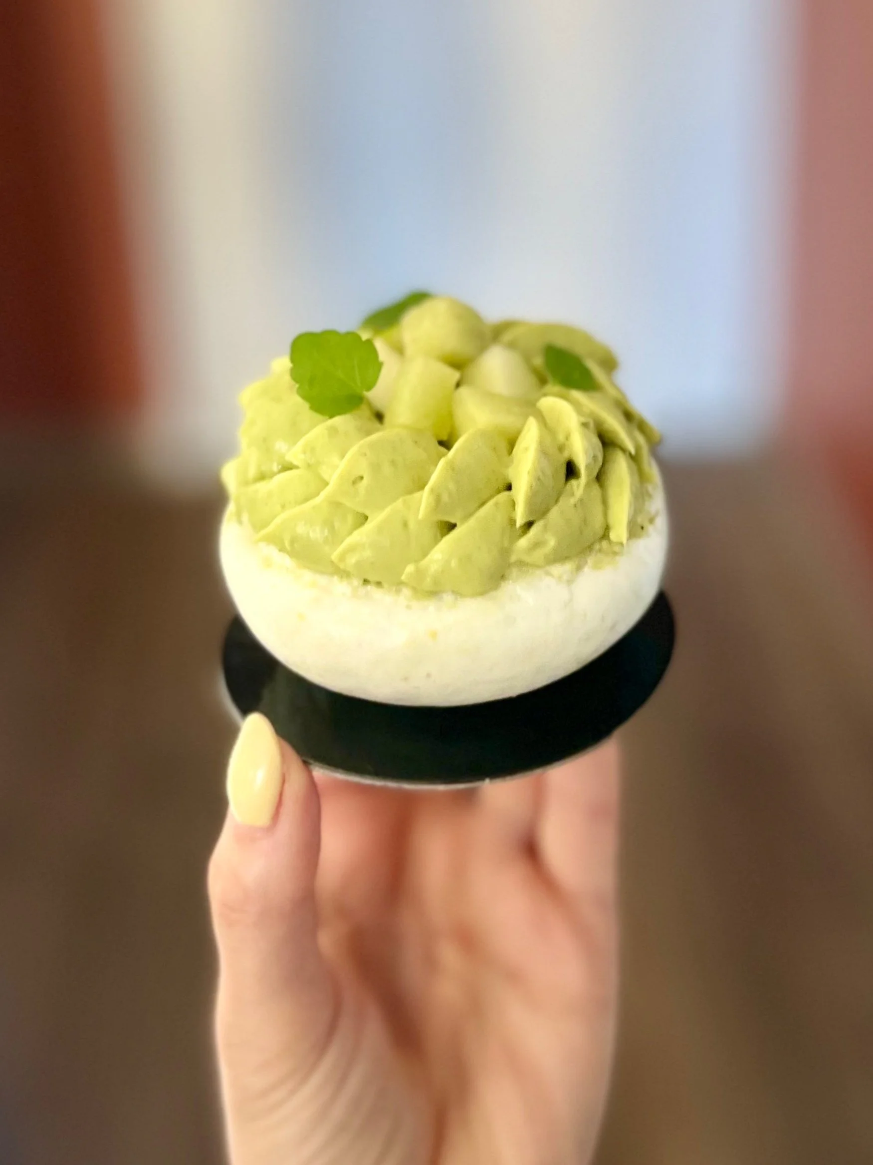 Gâteau décoré avec des touches vertes, garniture de pâte verte et petites feuilles vertes, transporté sur une petite assiette noire, tenu par une main avec ongle jaune.