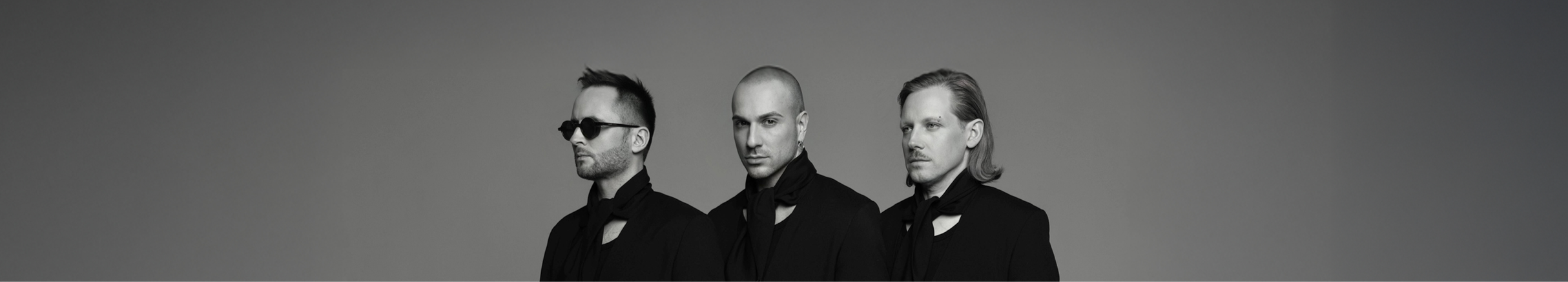Black and white photo of three men standing side by side, wearing black suits and dark clothing, against a plain gray background.