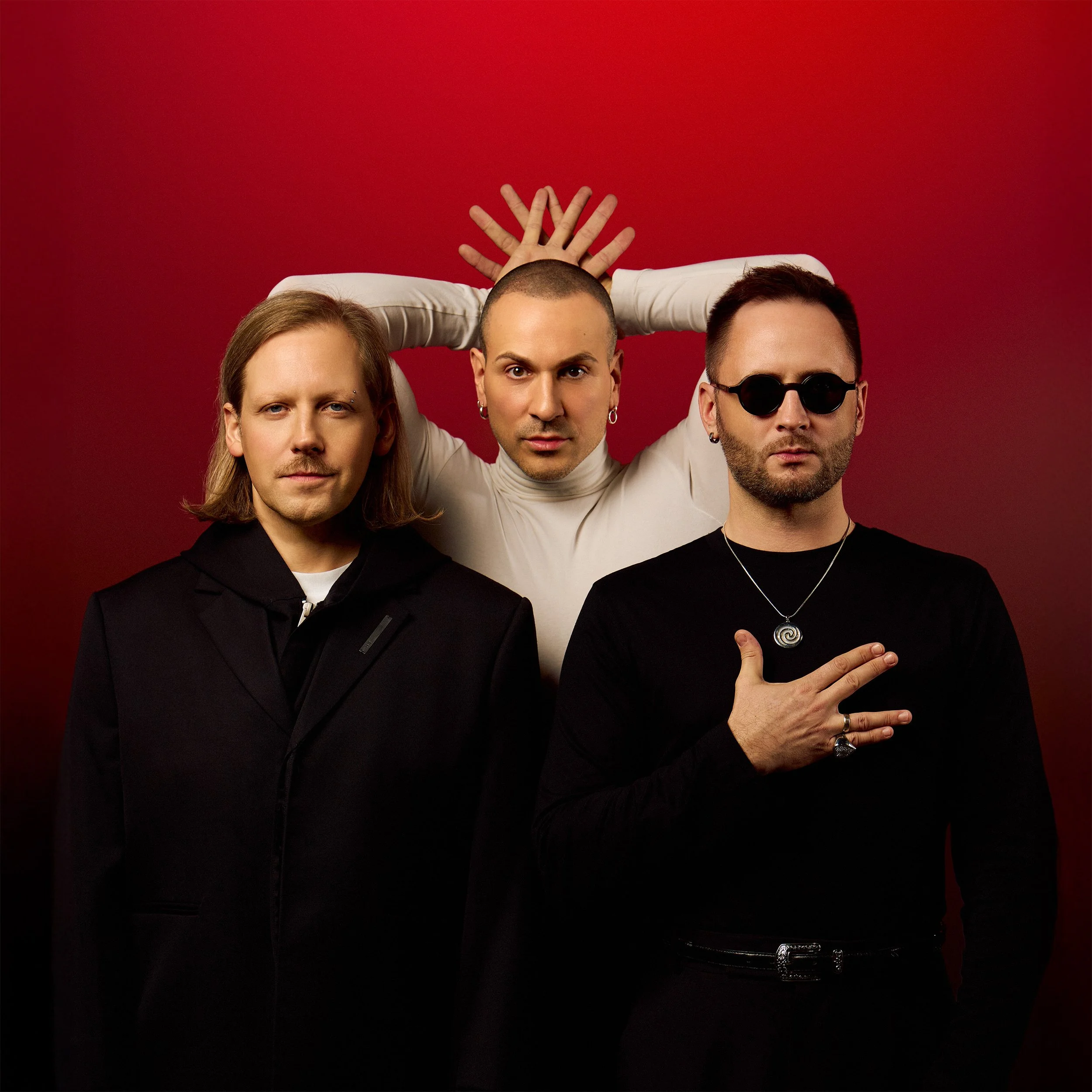 Three men posing against a red background, with the man in the middle standing behind with arms raised and hands behind head, the man on the left dressed in a black coat, and the man on the right wearing sunglasses and jewelry.