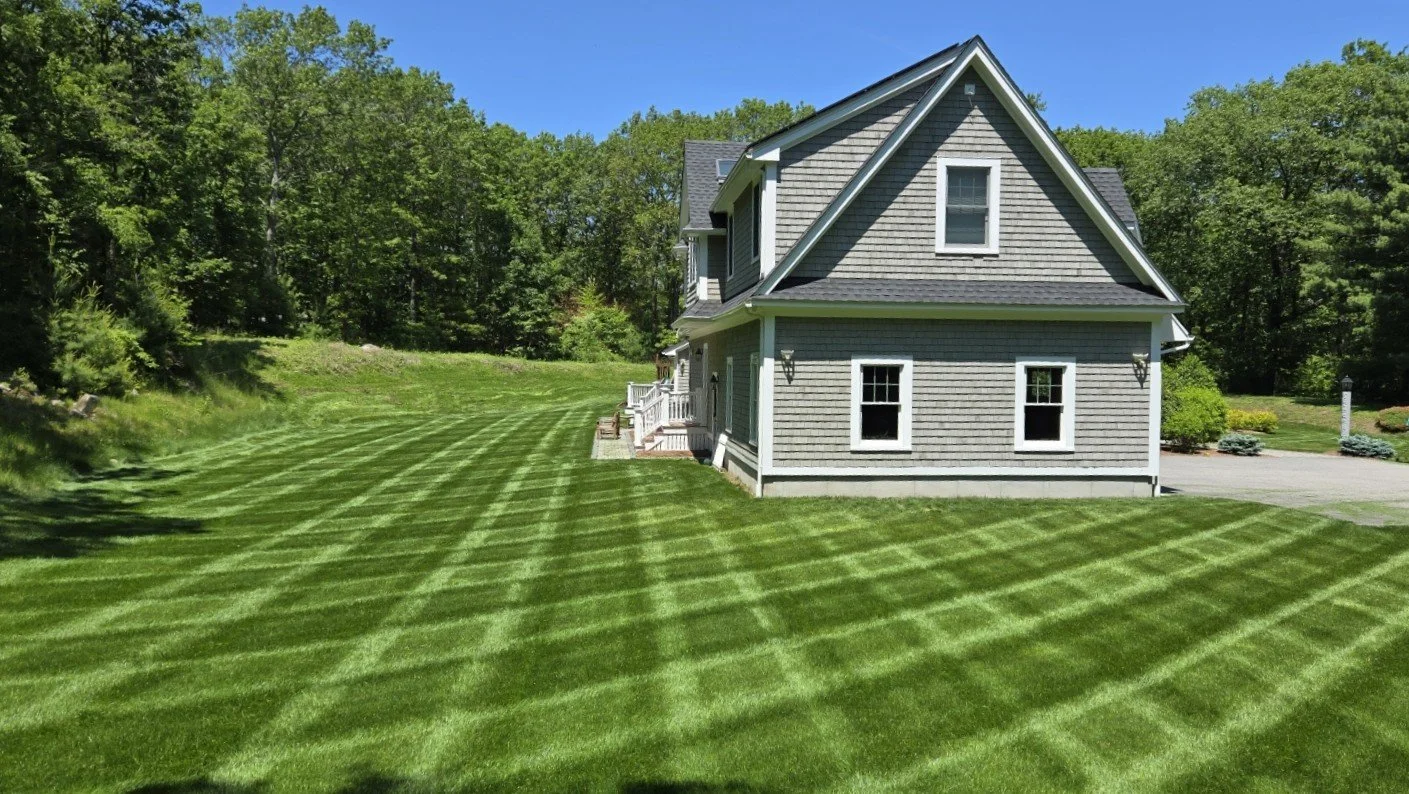 A house with grey siding and white trim, surrounded by a well-maintained green lawn with striped mowing pattern, some trees in the background, and a clear blue sky overhead.