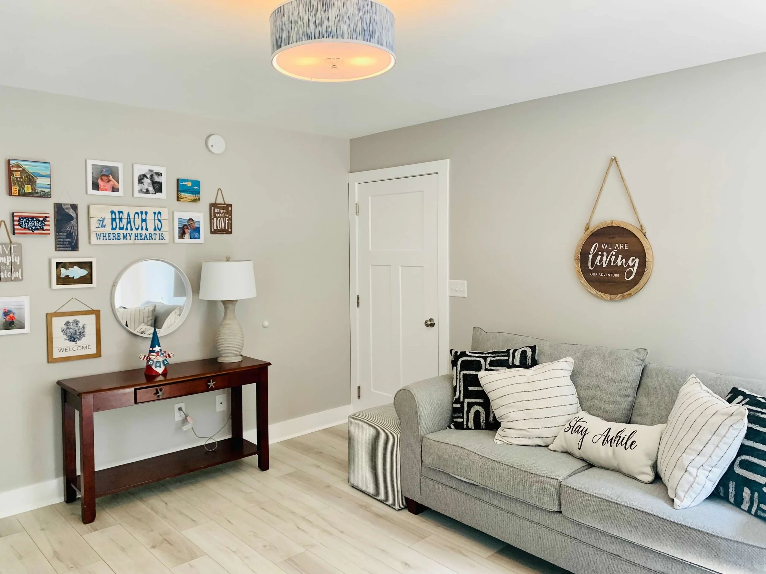 Cozy living room with light-colored walls, a gray sofa with decorative pillows, a wooden console table with a lamp and wall art above, and a hanging round sign that says "We Are Living Our Adventure".