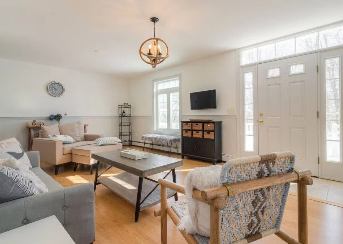 Living room with beige sofas, wooden coffee table, chair with woven fabric, wall-mounted TV, large front door with sidelights, window, and hardwood floors.