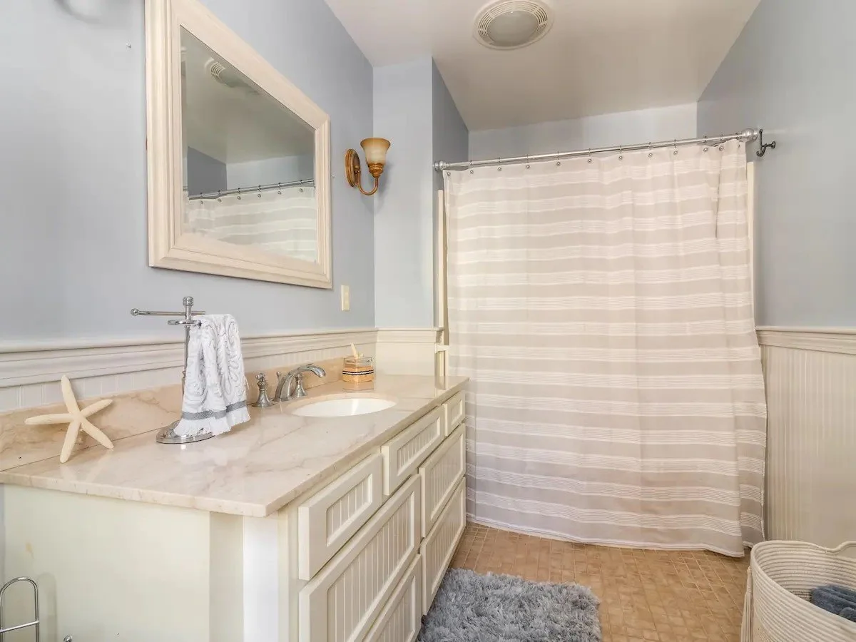 Bathroom with light blue walls, a large mirror, a white vanity with a beige marble countertop, a starfish decorative item, a towel, a basket, a shower curtain, and a gray rug.
