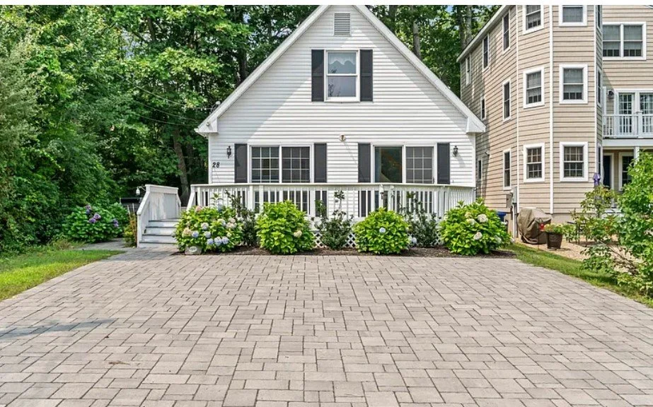 White house with black shutters and a small front porch, surrounded by greenery and a paved driveway.