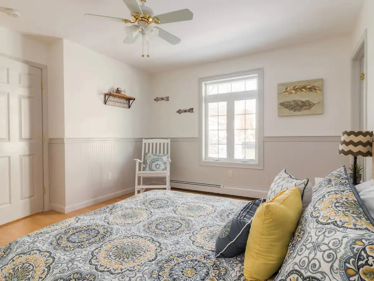 Bedroom with white walls, wooden floor, bed with patterned bedding and yellow and gray pillows, window allowing natural light, white chair, wall decor including paintings and fish-shaped decorations, ceiling fan with white blades and gold accents, with a lamp on nightstand.