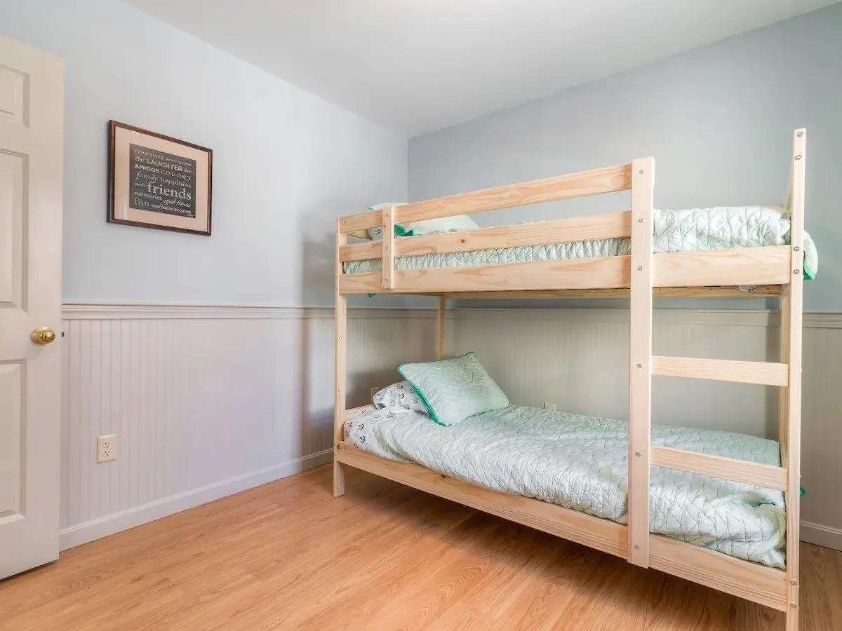 A bedroom with light blue walls, showing a wooden bunk bed with light green bedding and pillows. There is a framed poster on the wall and a wooden floor.