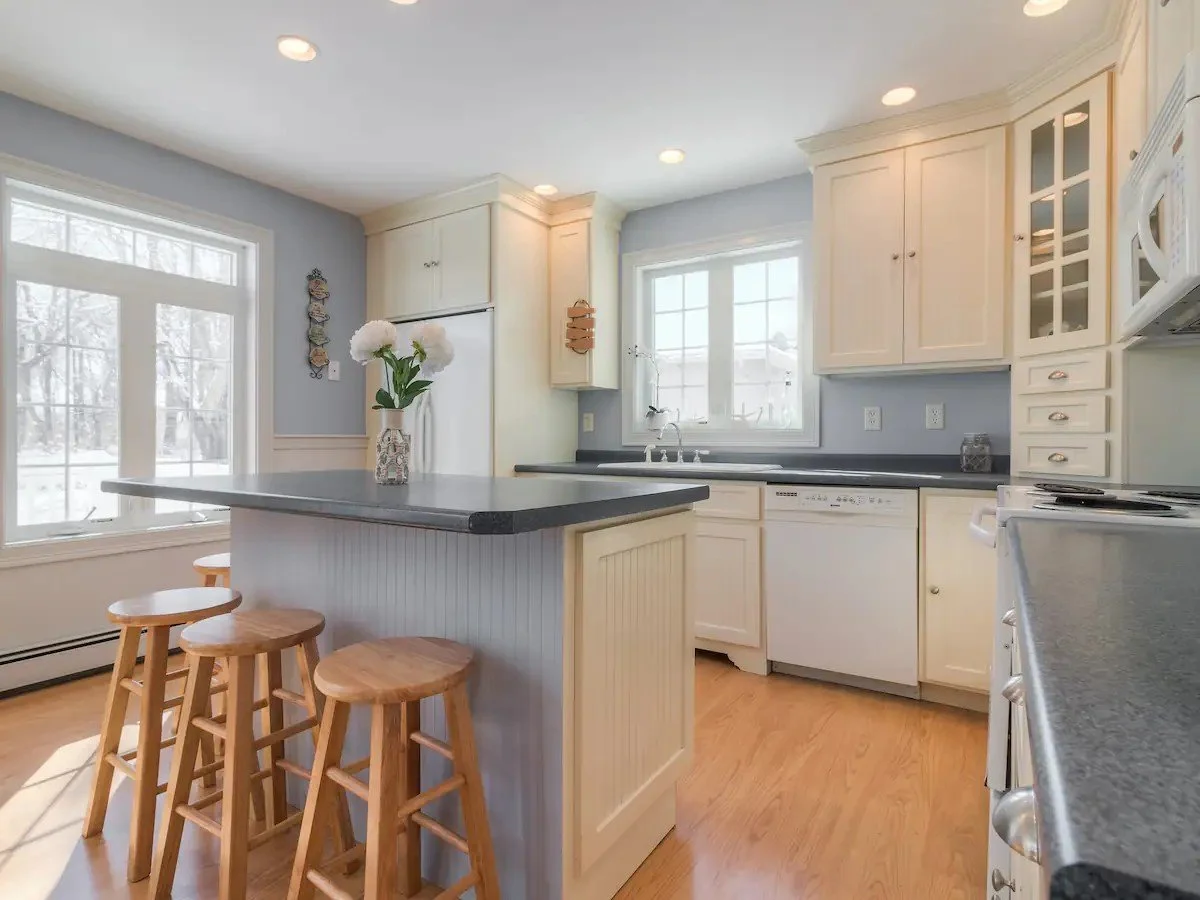 Bright kitchen with white cabinets, black countertops, a central island with a vase of white flowers, bar stools, and large windows allowing natural light.