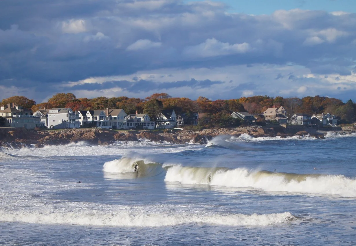 A coastal scene with waves crashing onto the shore and surfer riding a wave, with houses on a rocky cliff in the background under a cloudy sky.