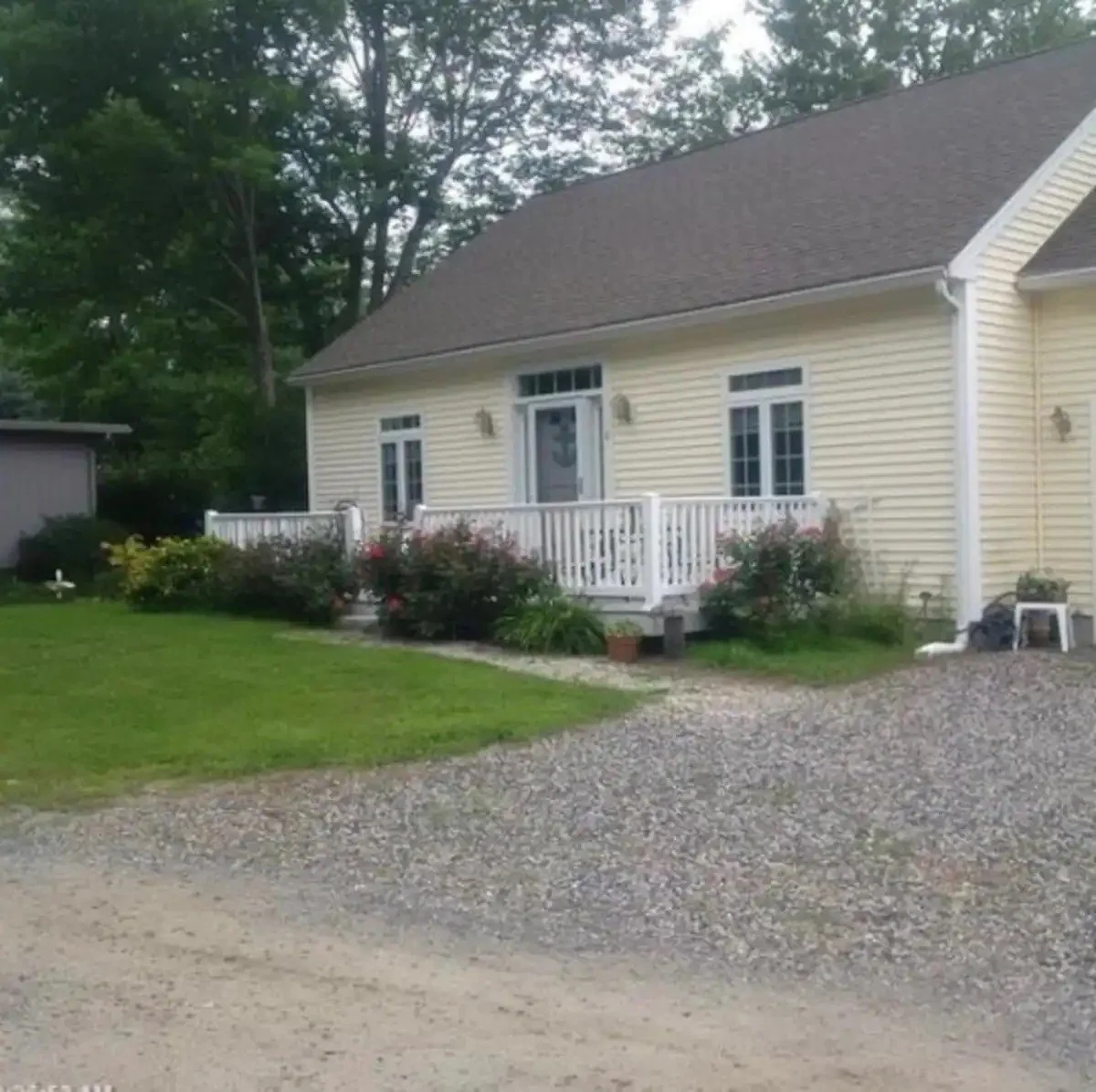 A yellow house with white trim and a small porch with railing, surrounded by bushes and flowers, with a gravel driveway in front.