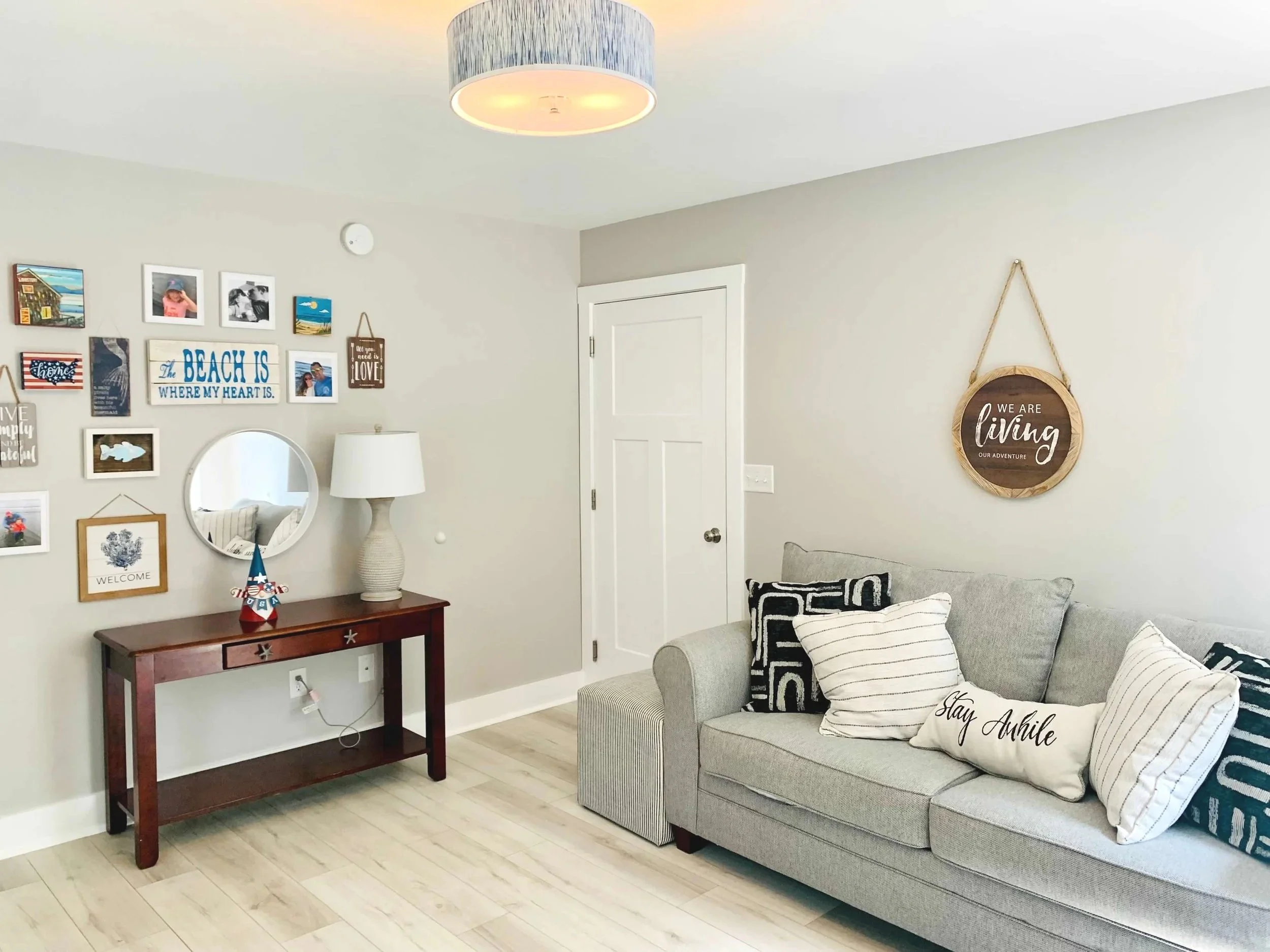 A living room corner with a light grey sofa, patterned pillows, a wall decor sign that says "We are living our adventure," a wooden table with a decorative item, a mirror, and a gallery wall with various framed pictures and signs. A white lamp is placed on the table, and the room has light-colored wood flooring and a white door.