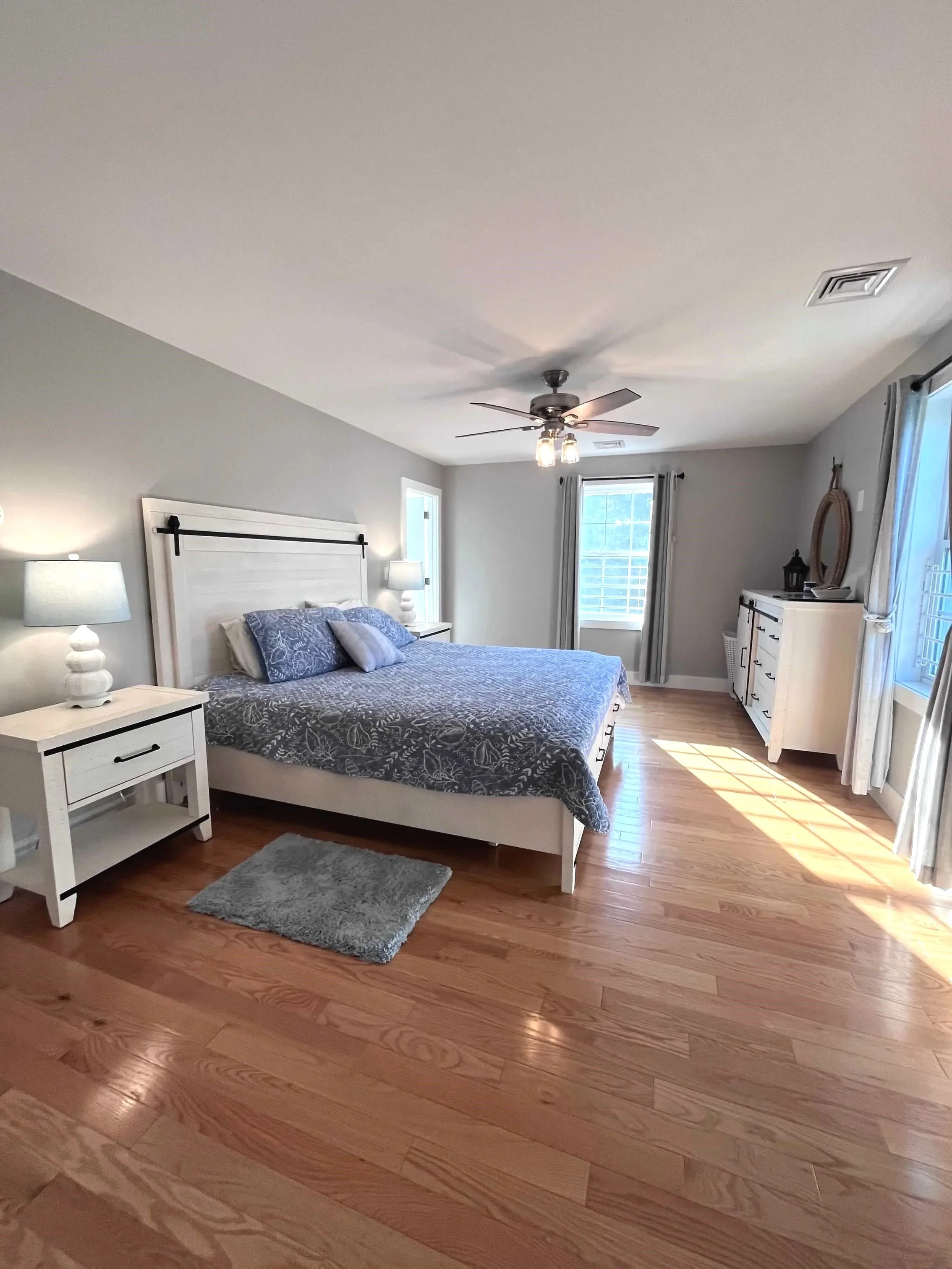 A bedroom with a white bed, blue patterned linens and pillows, two bedside lamps, a light-colored dresser, a ceiling fan with lights, hardwood floors, and windows with curtains.