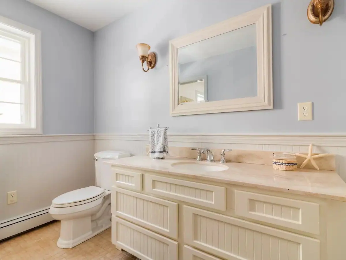 Bathroom with a white vanity, beige countertop, large wall mirror, wall-mounted light fixtures, toilet, window, starfish decor, and wood flooring.