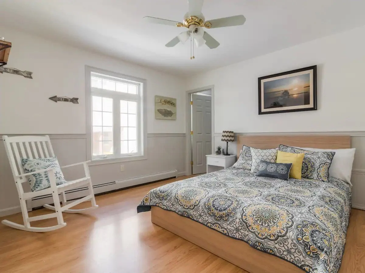 Bedroom with a rocking chair, a window, a bed with patterned bedding and multiple pillows, a nightstand with a lamp, and framed artwork on the walls.