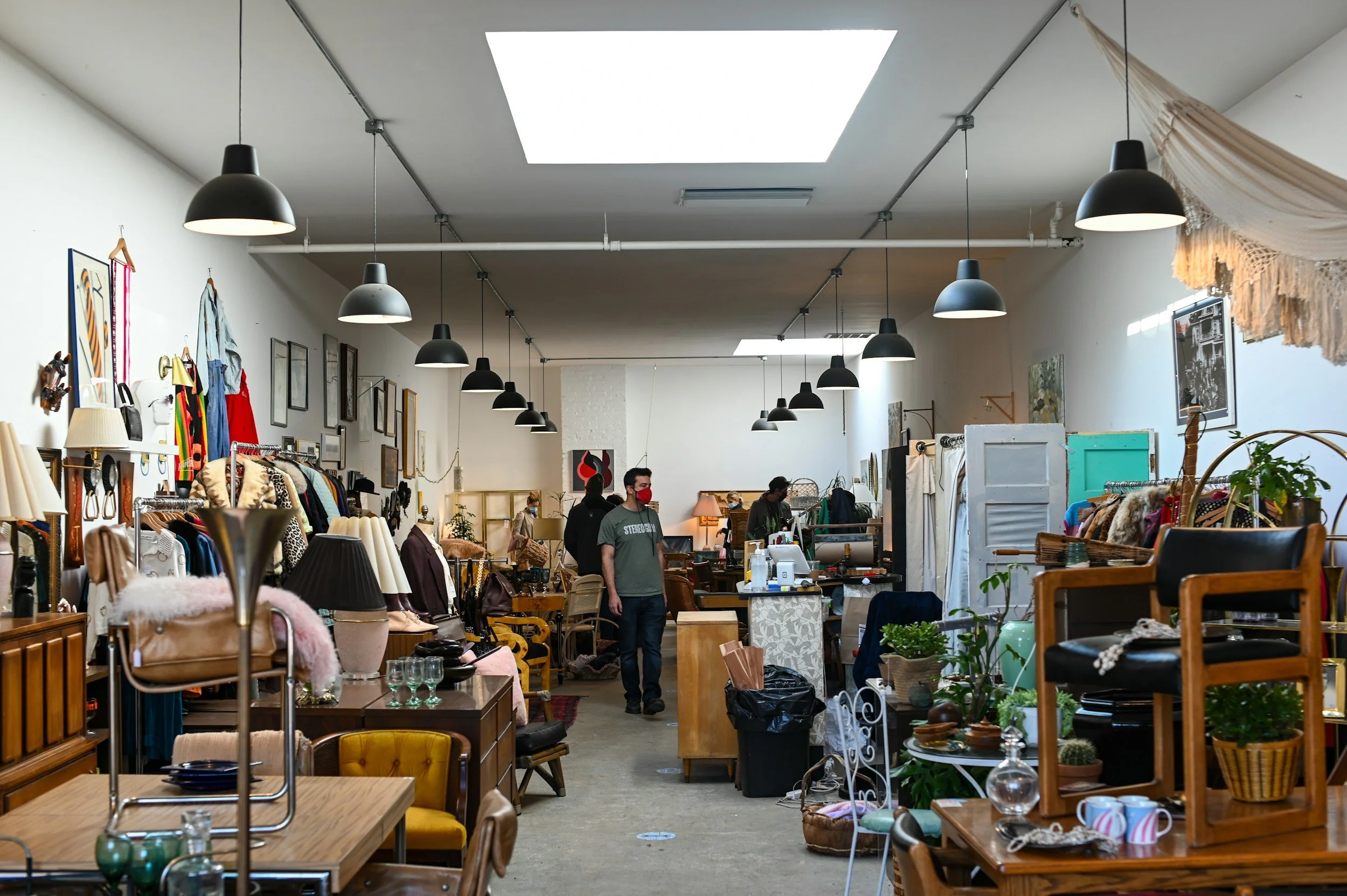 Interior of a thrift store with vintage furniture, clothes, lamps, and decorative items, with several people shopping and browsing.