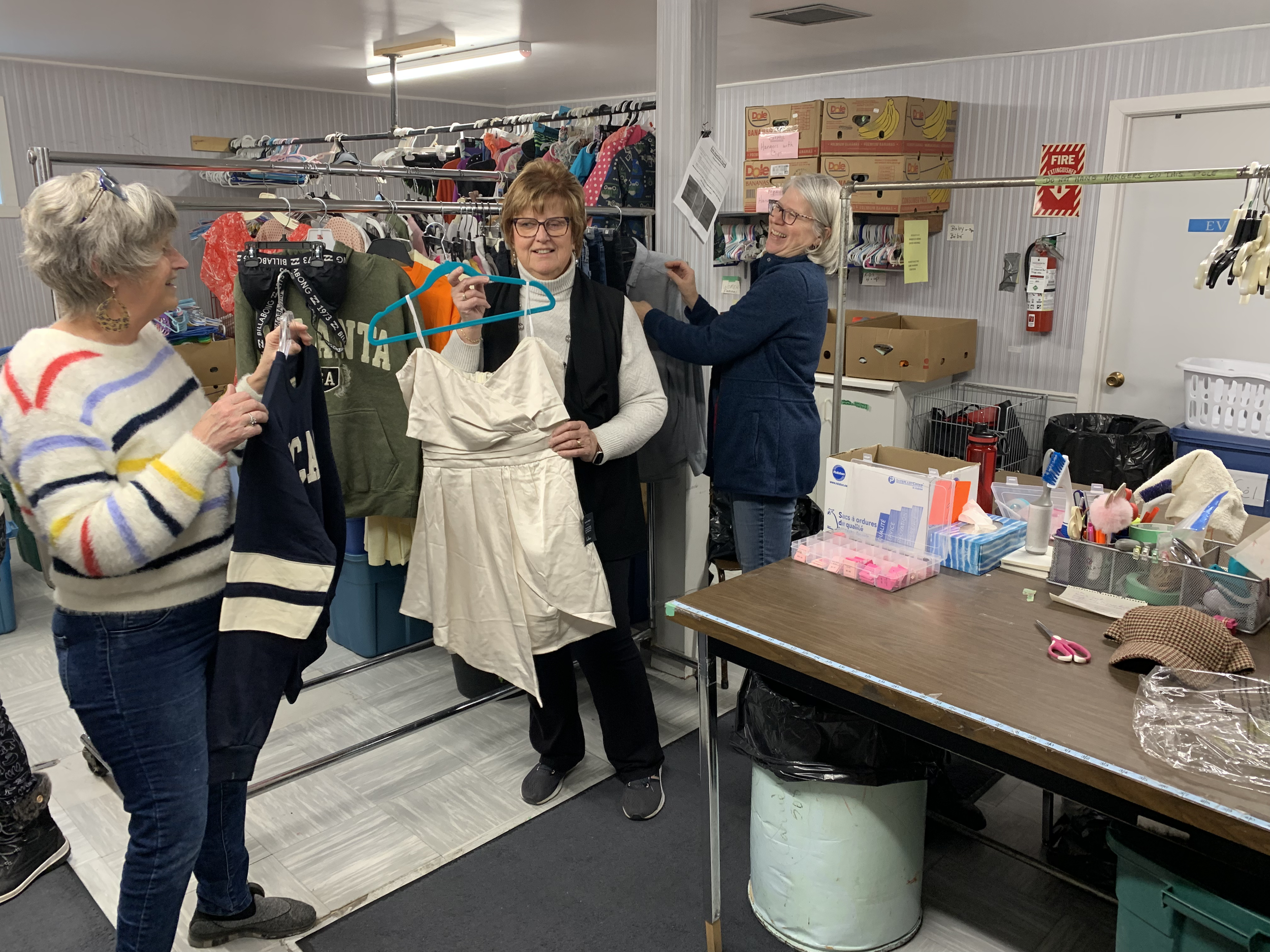 Three women shopping and talking inside a thrift store, with clothing racks, boxes, and a table with various items around them.