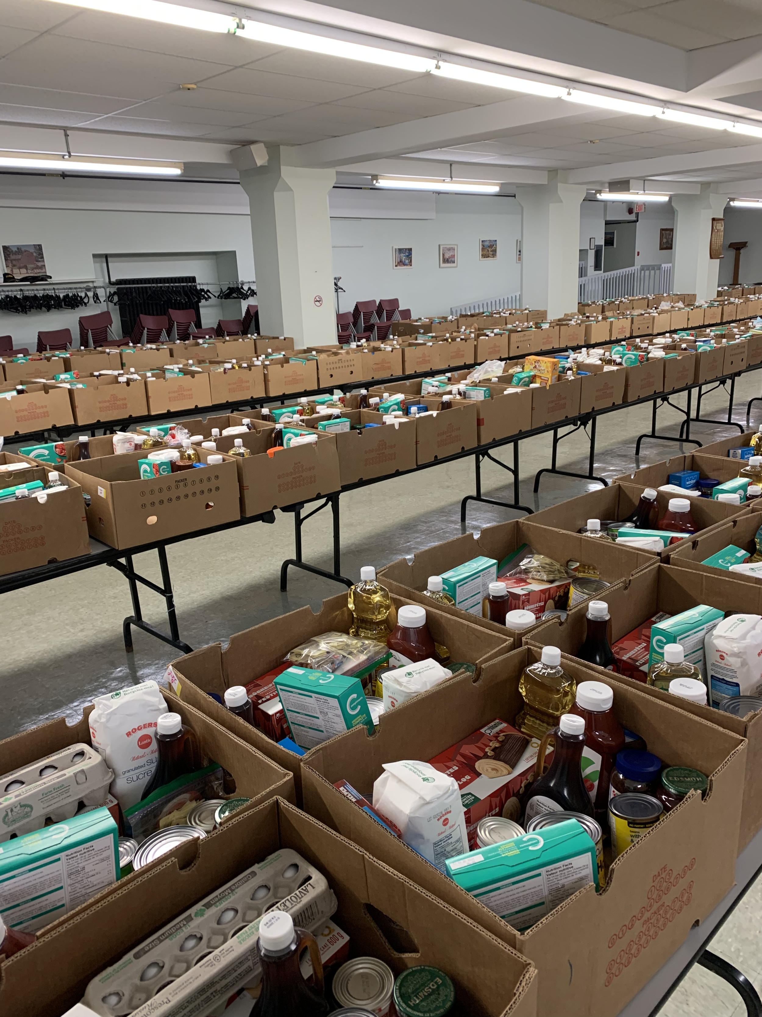 Multiple tables filled with boxes of non-perishable food items, likely organized for a community food drive or distribution event, in a large indoor space with chairs and artwork on the walls.