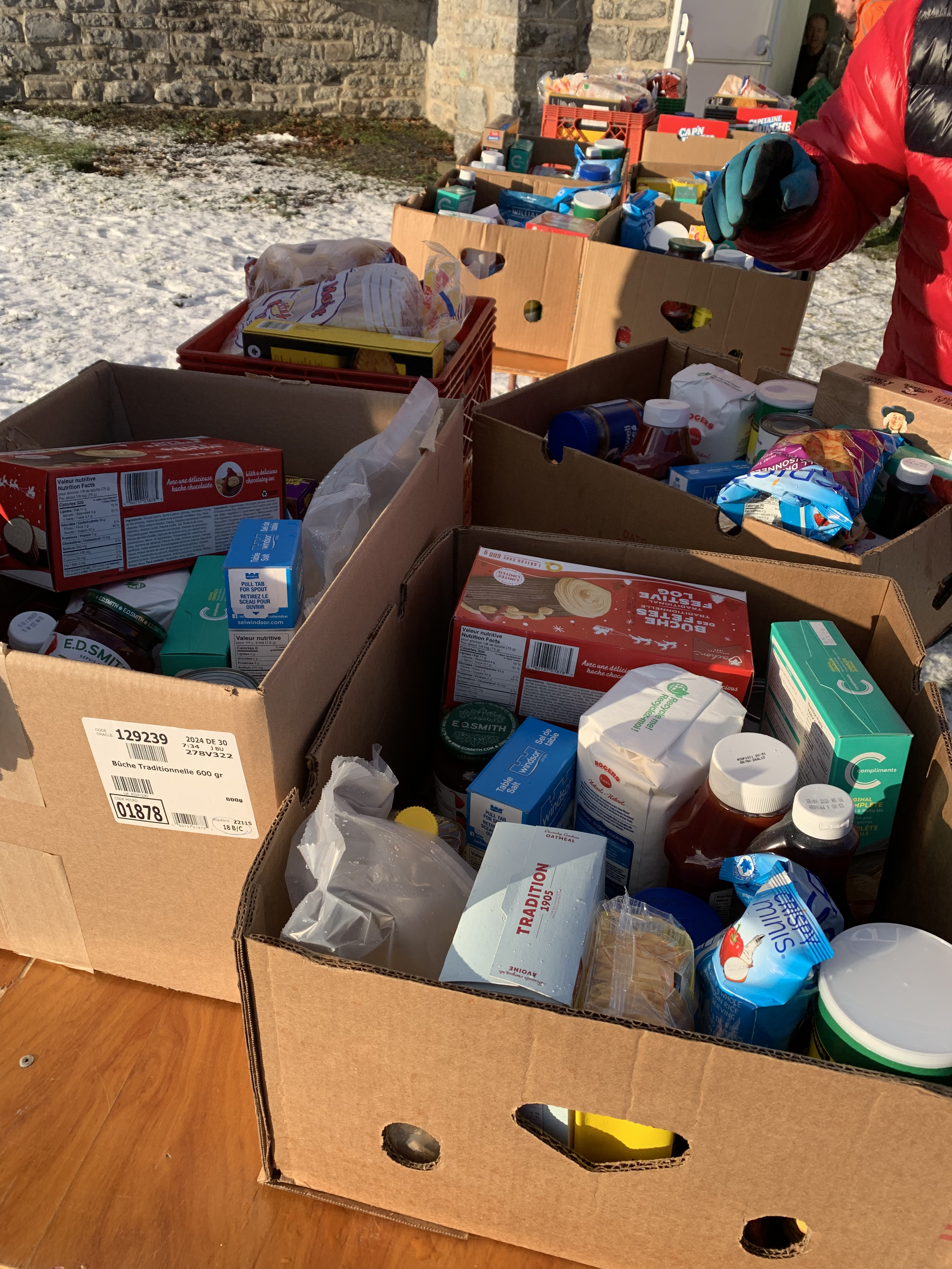 Multiple open cardboard boxes filled with grocery items on a wooden table outdoors, with snow on the ground and a stone wall in the background.