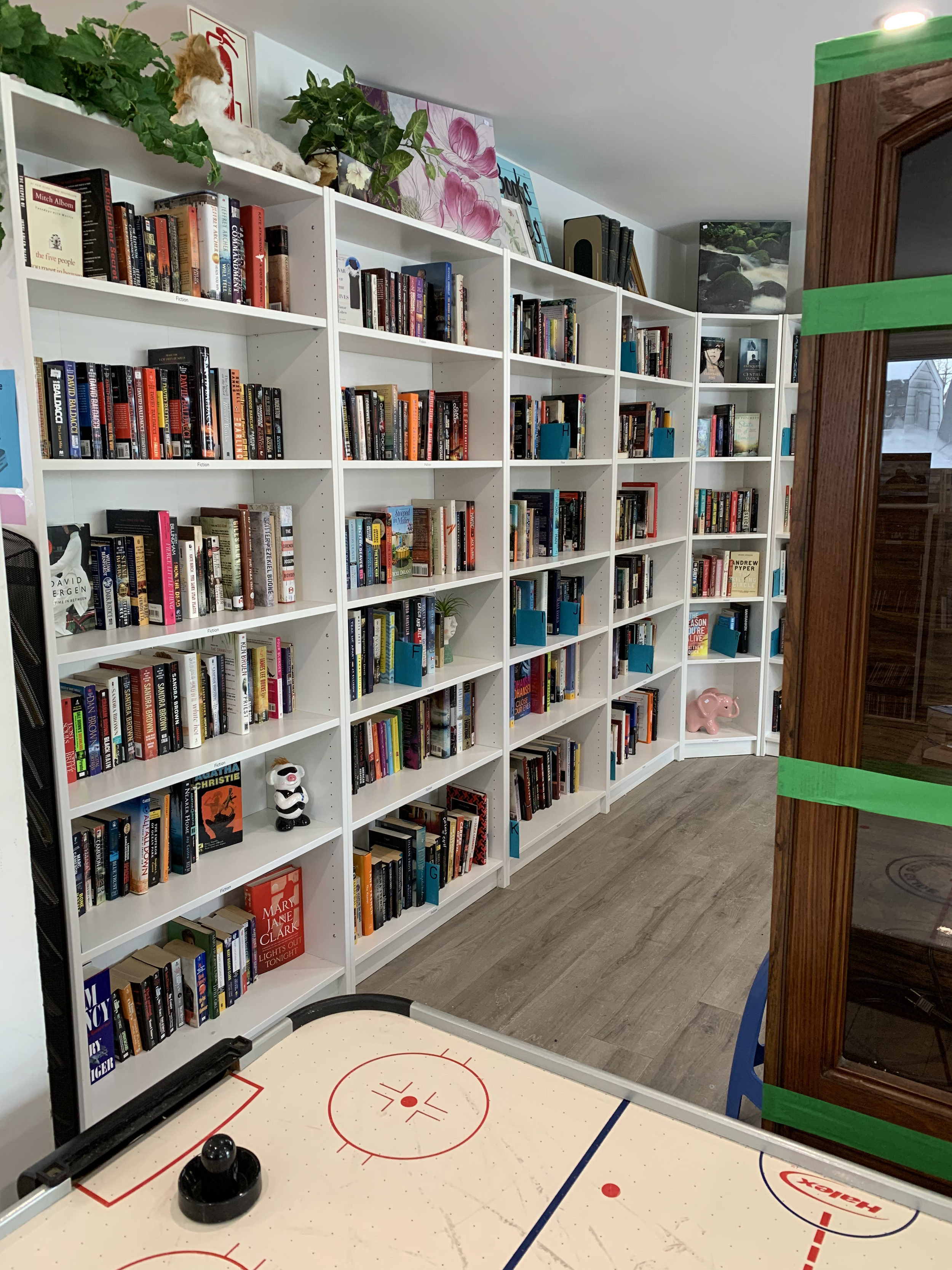 View of a bookstore with white bookshelves filled with various books and decorations, an air hockey table in the foreground, and a wooden display case with green tape on the right side.