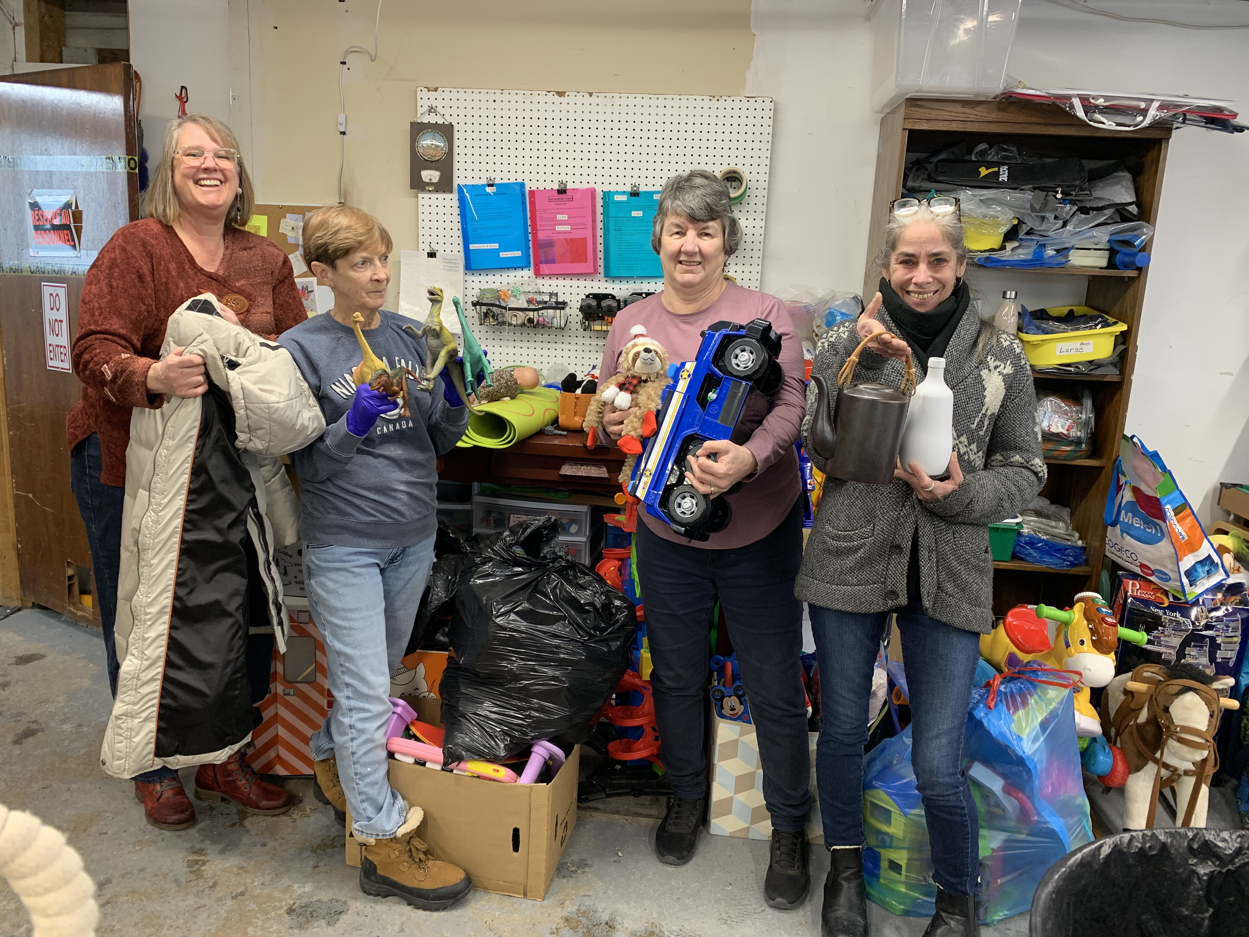 Four women standing in a room filled with toys and household items, smiling at the camera.