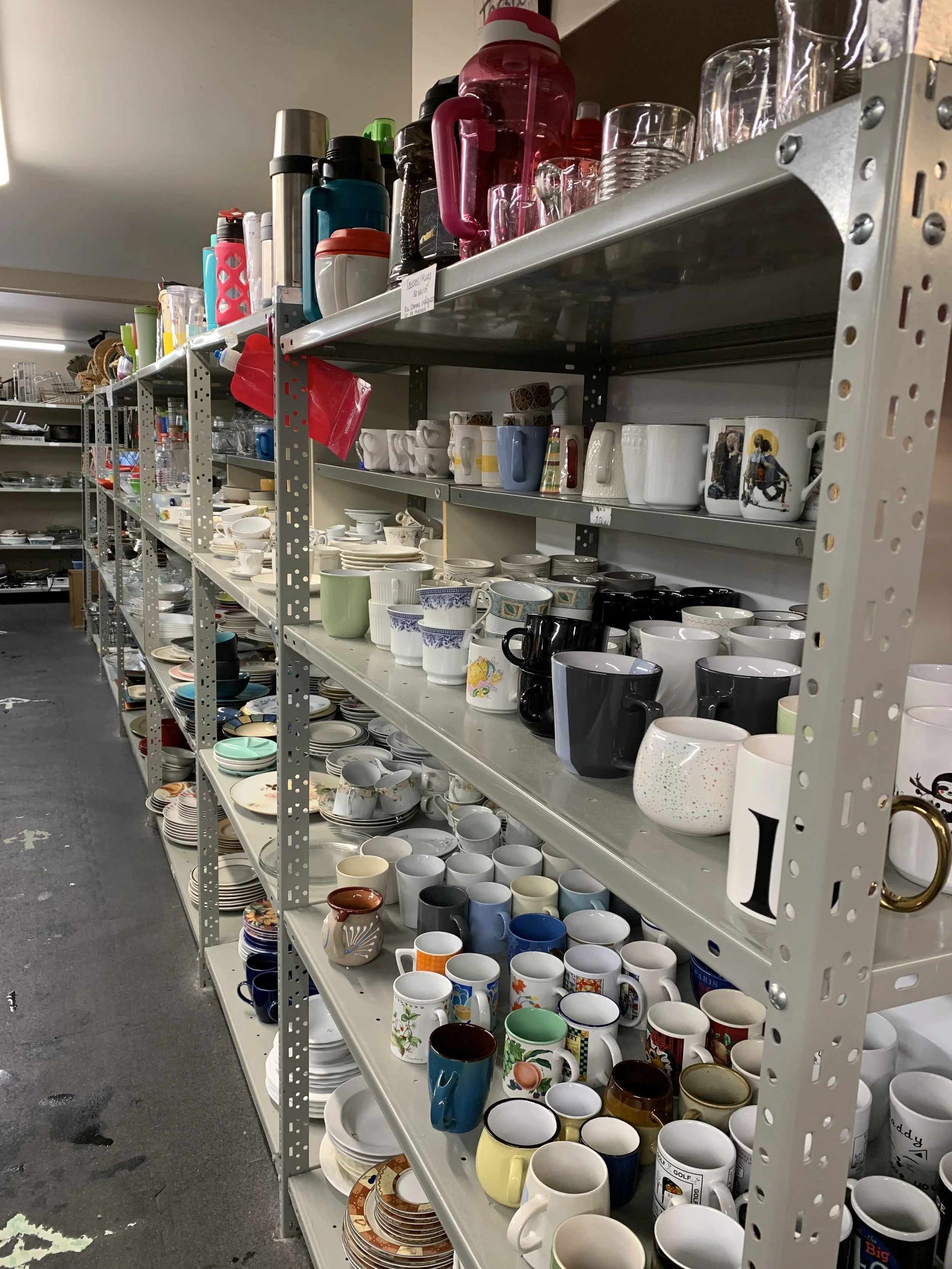Metal shelving units filled with various mugs, cups, and glassware in a thrift store or warehouse setting.