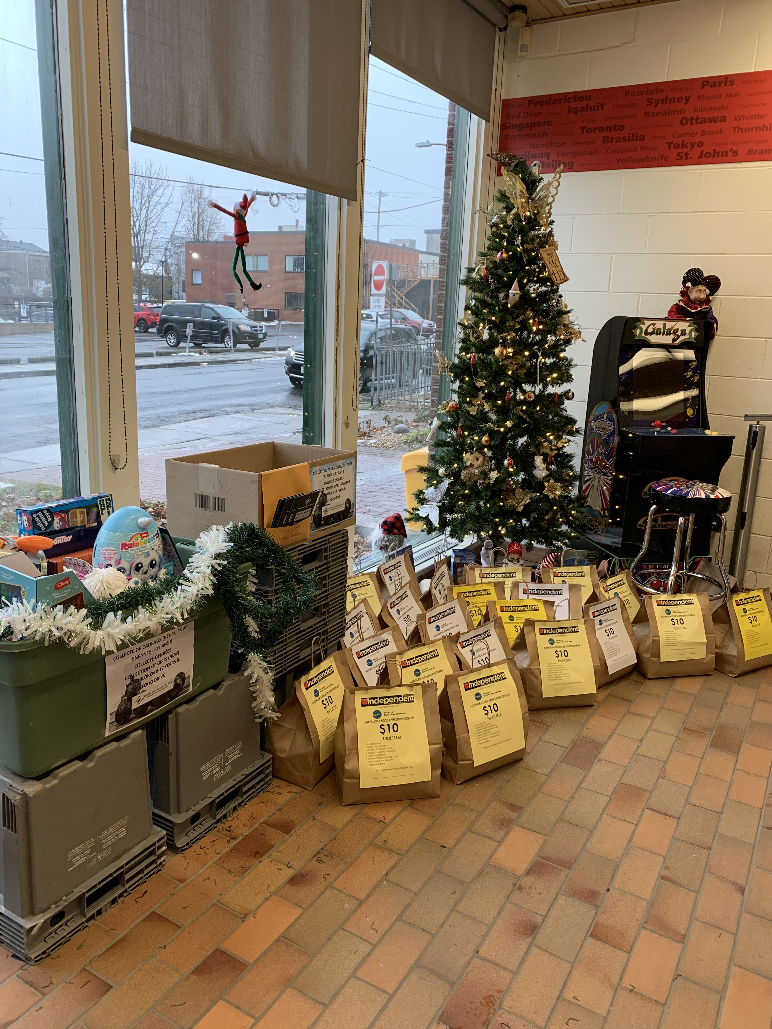 Indoor scene with a decorated Christmas tree, donations bags labeled "Independent," and a vintage pinball machine, near large windows showing a wet street and parked cars outside.