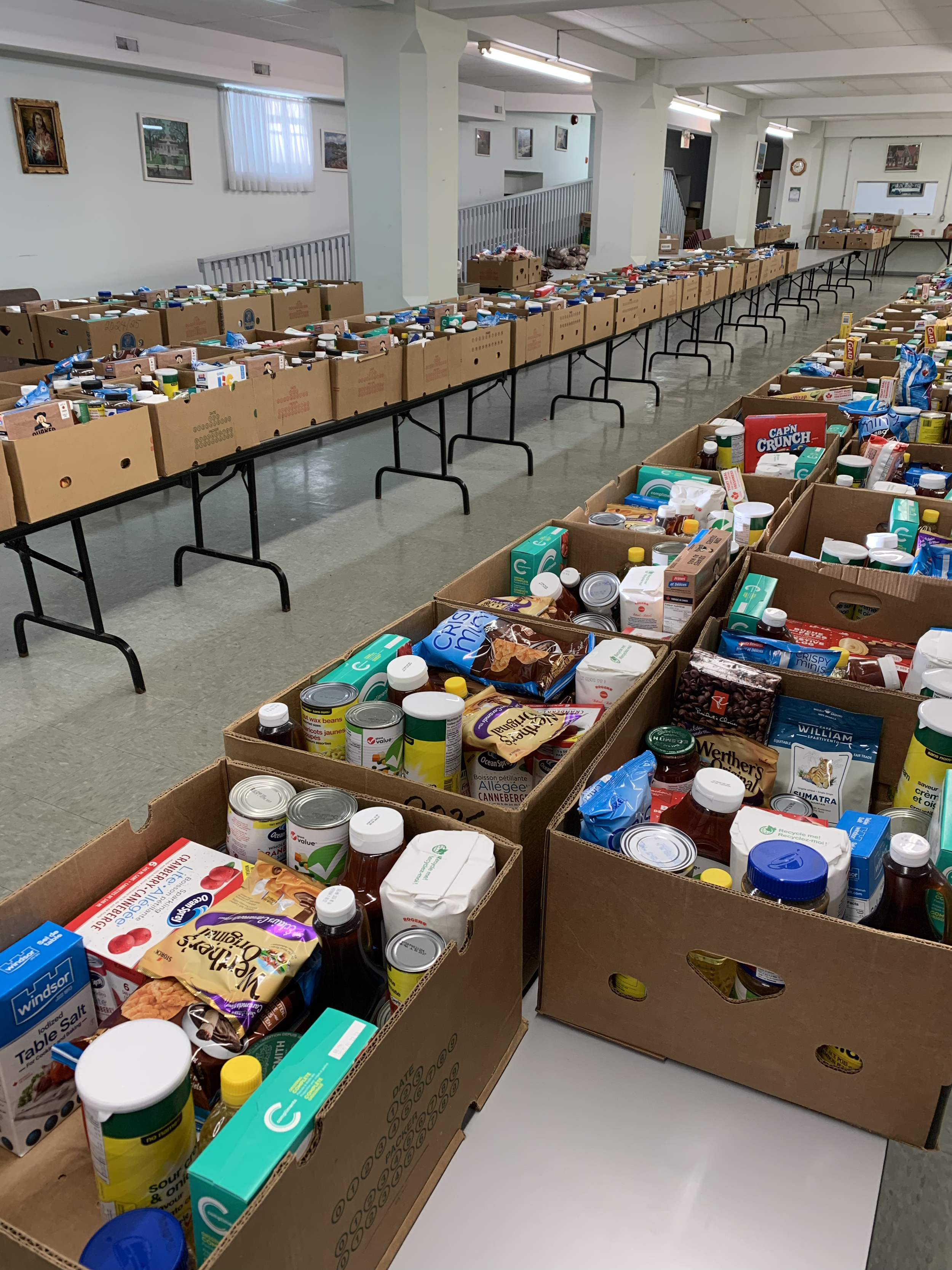 Long rows of cardboard boxes filled with groceries on tables in a community center or similar venue.