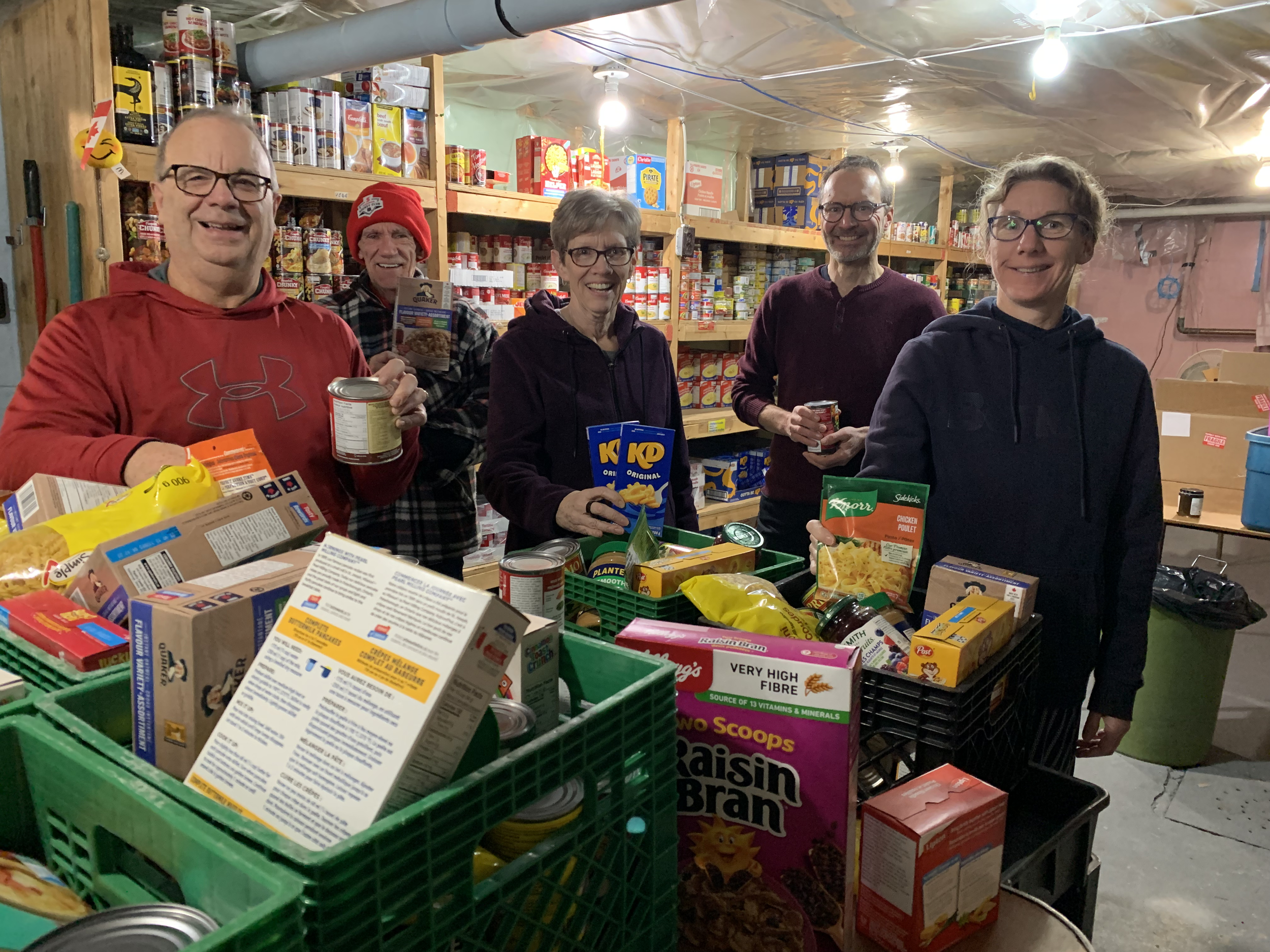 Six people standing behind a table of canned and boxed food in a storage room with shelves of canned goods in the background.