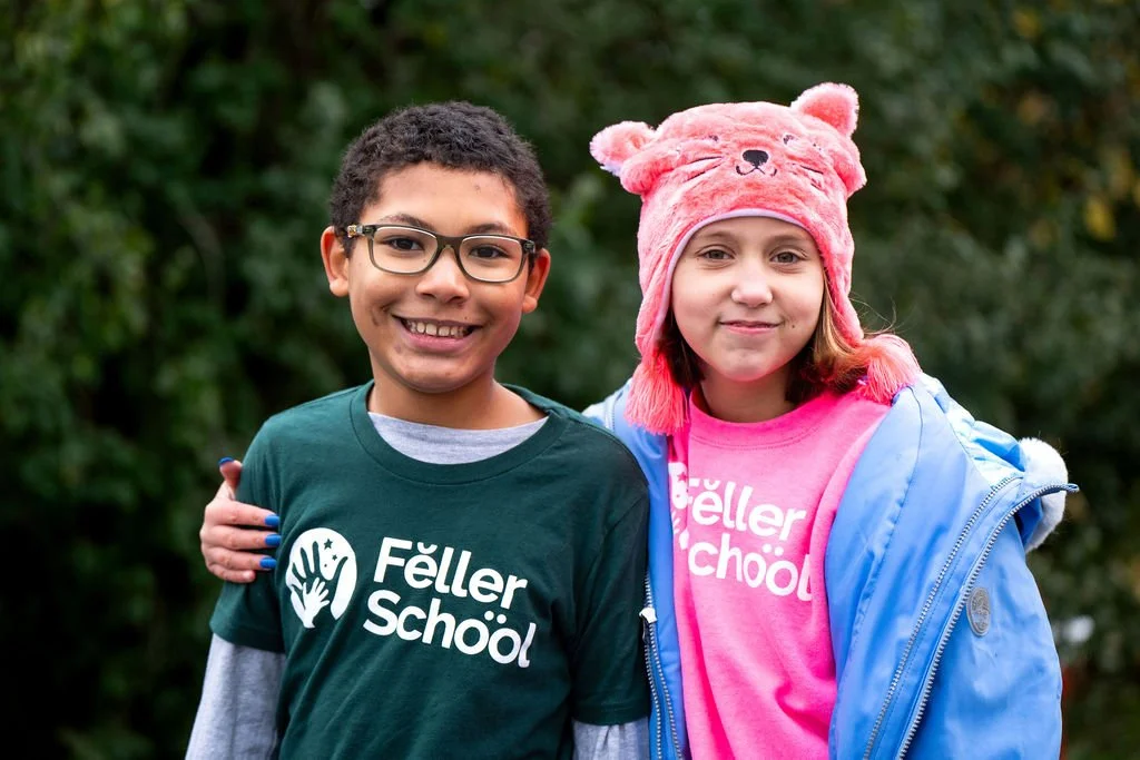 Two children smiling outdoors, one with glasses wearing a dark green Feller School t-shirt and the other in a pink Feller School t-shirt with a pink bear hat and a blue jacket.