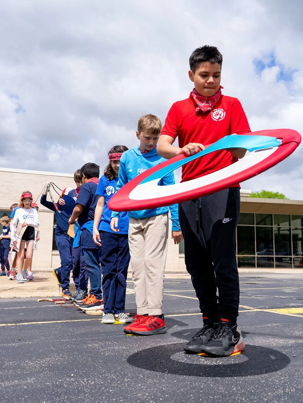 Group of children standing in line on a schoolyard, playing with a hula hoop and jump rope, under a cloudy sky.