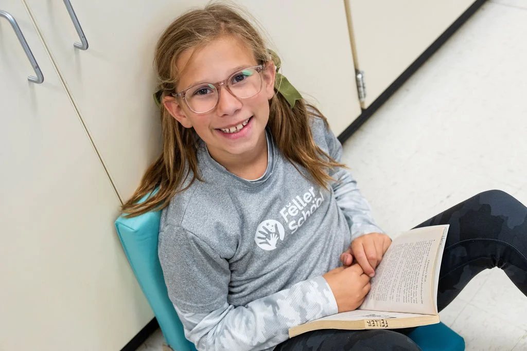 A smiling girl with glasses sitting on the floor, leaning against a wall, holding a book, wearing a gray 'Fletcher School' T-shirt.