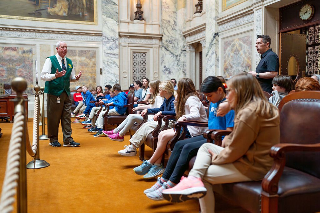 A guide in a green vest and white shirt is speaking to a group of children and adults seated in chairs inside a grand, marble-walled hall with a clock and ornate decorations.