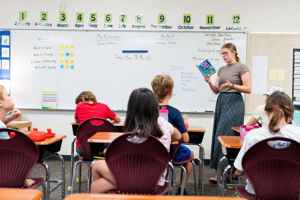 Elementary school classroom with students seated at desks and a teacher reading a book to them. The whiteboard behind displays a calendar with months numbered and names written. Students are focused on the teacher, who is standing by the whiteboard, wearing a gray shirt and patterned skirt.